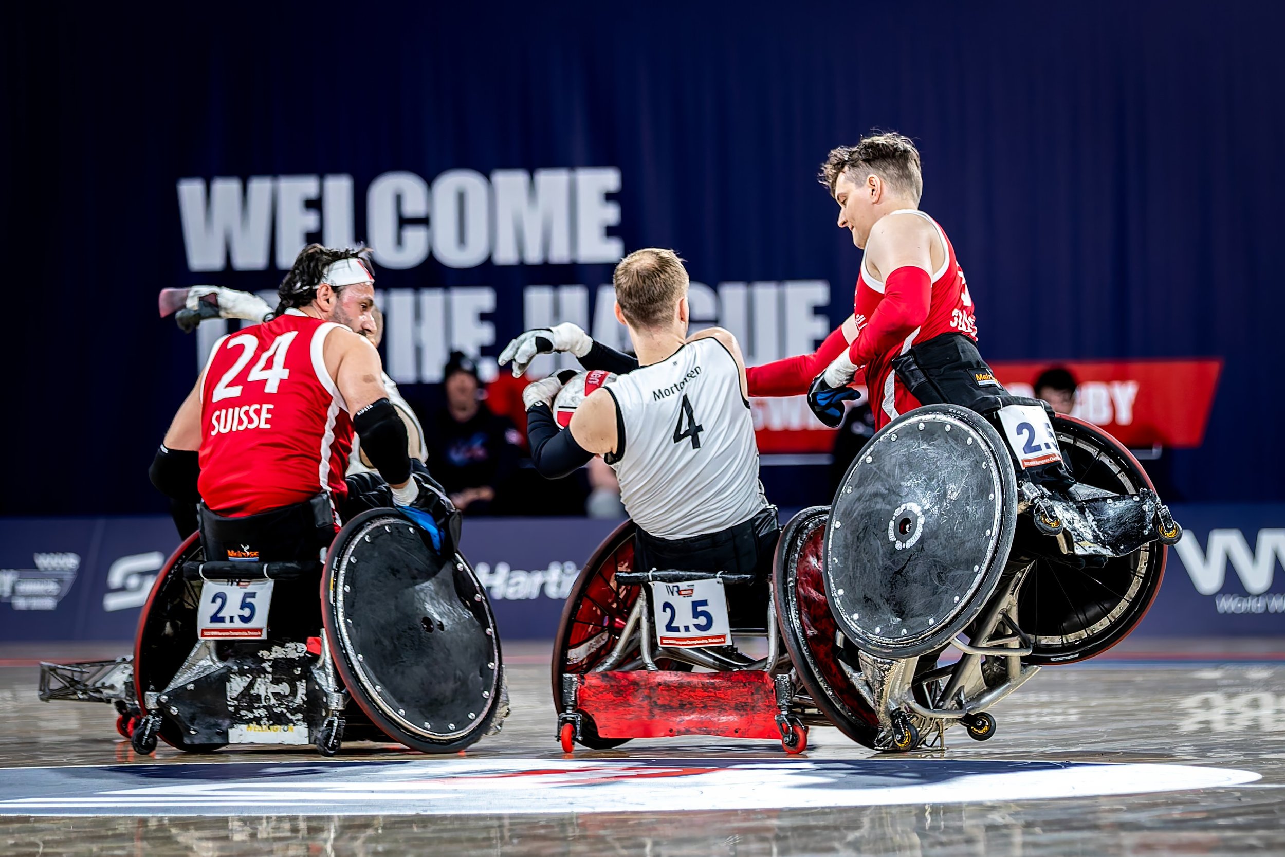 Wheelchair rugby players in red and white jerseys celebrating on the court with a large sign in the background that reads 'WELCOME'.