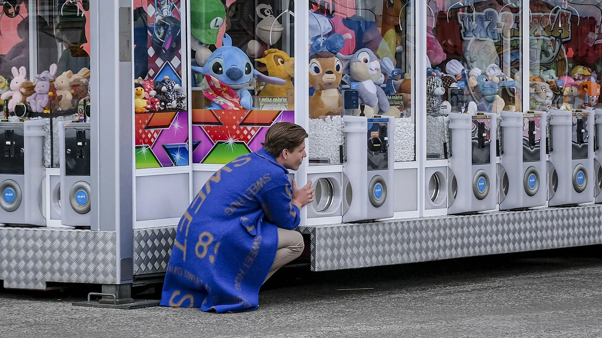 A young man crouches in front of a carnival claw machine filled with plush toys, including characters from animated movies, with colorful game displays and coin slots.