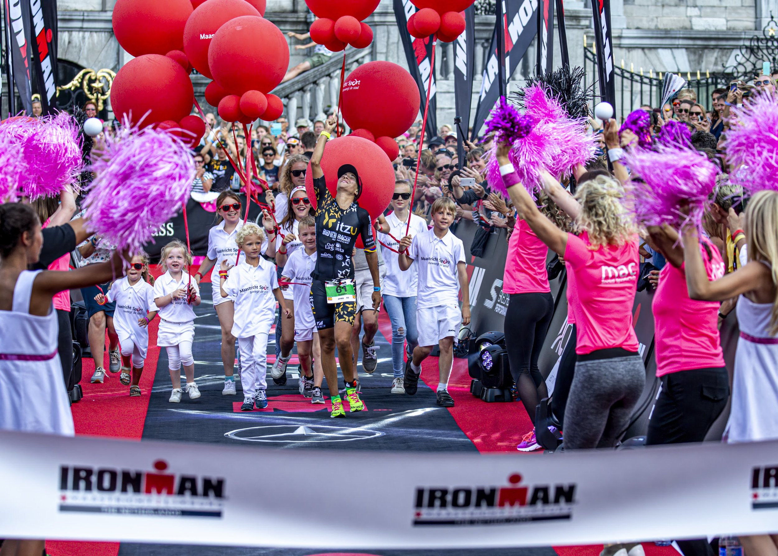 Participants crossing the finish line at an Ironman race event, surrounded by spectators and event staff, with large red balloons and pink feather boas in the scene.