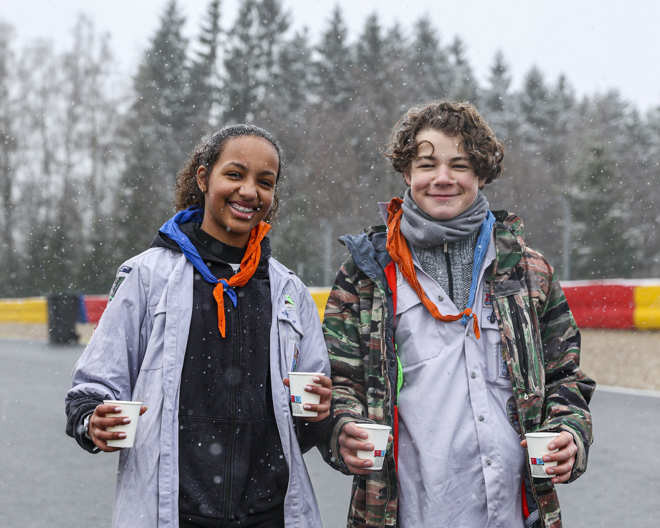 Two children, a girl and a boy, standing outdoors in winter, holding cups, with snow falling around them, and trees in the background.