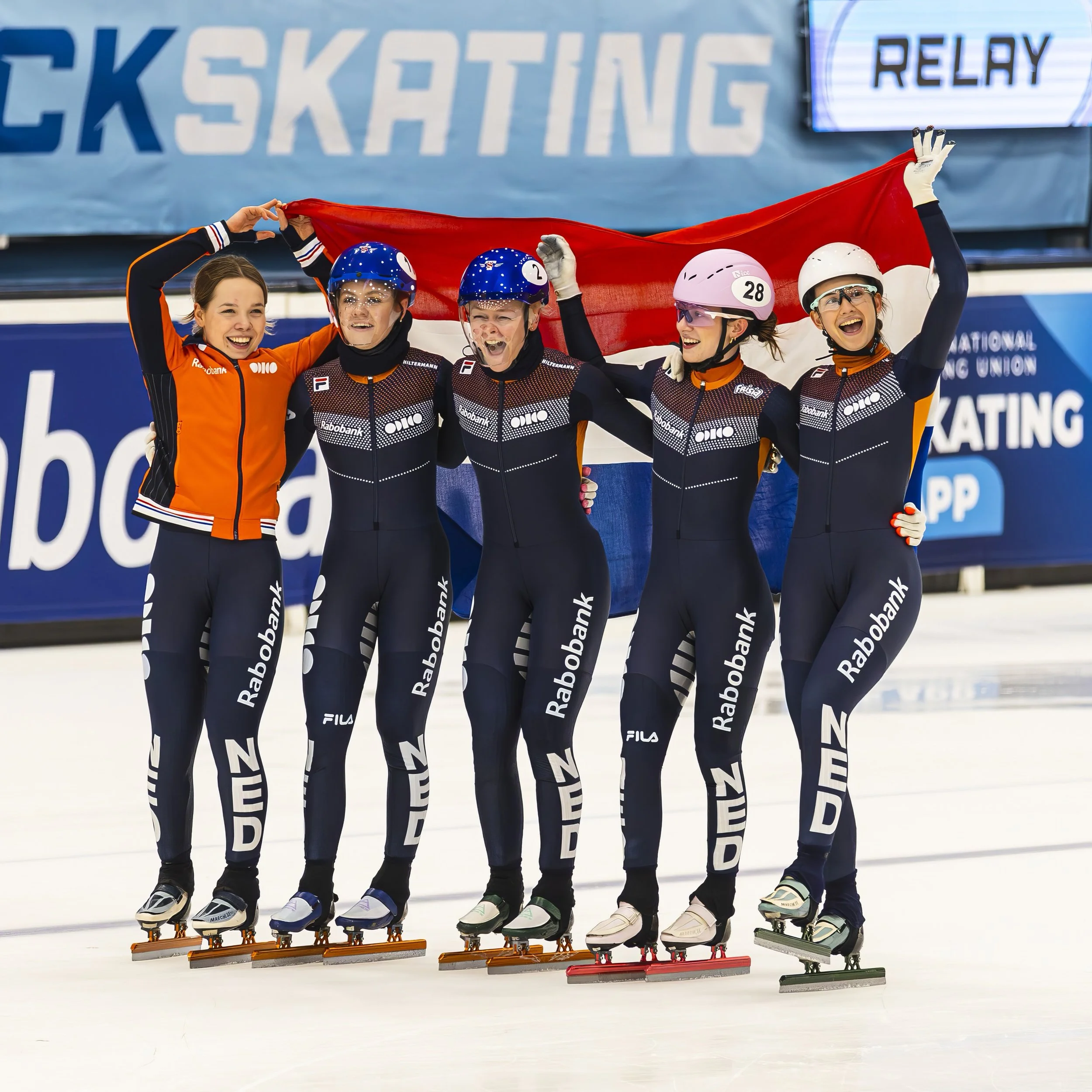 A group of five female ice skaters in uniform celebrating and holding a flag behind them on an ice rink, with banners and symbols indicating a competitive event.