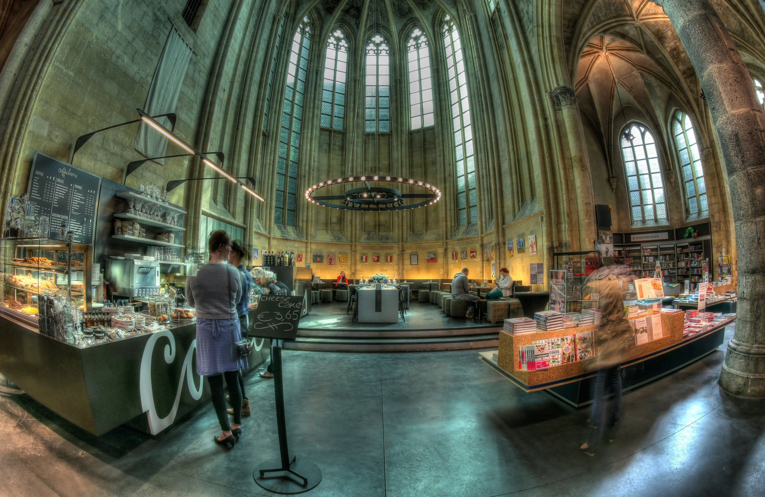 Interior of a large, cathedral-like building with tall stained glass windows, high vaulted ceilings, and stone columns. There is a cafe area with a counter, people seated at tables, and a bookshop with shelves of books and display tables.