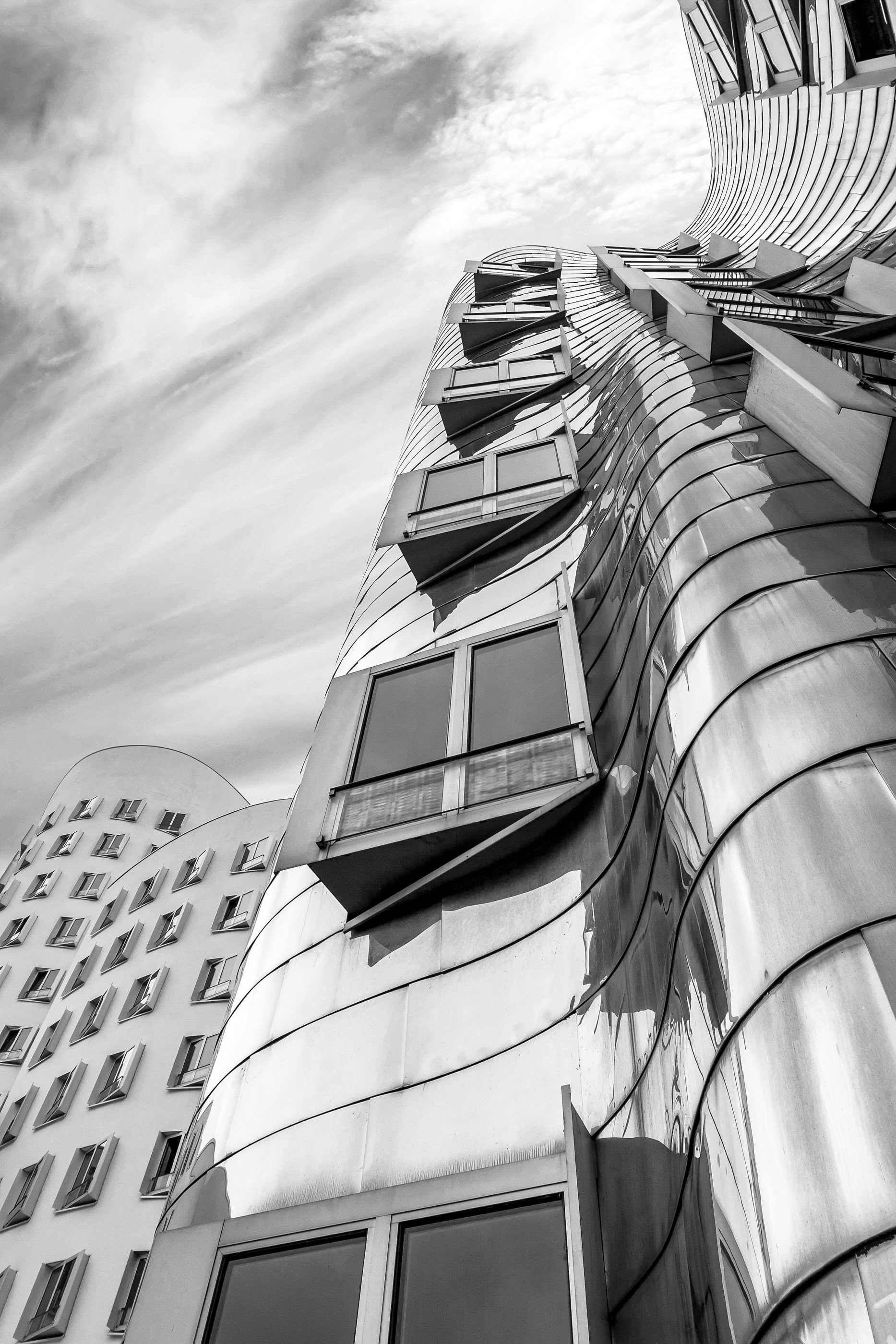 Black and white photo of modern, curved building with multiple windows and balconies, taken from a low angle against a cloudy sky.