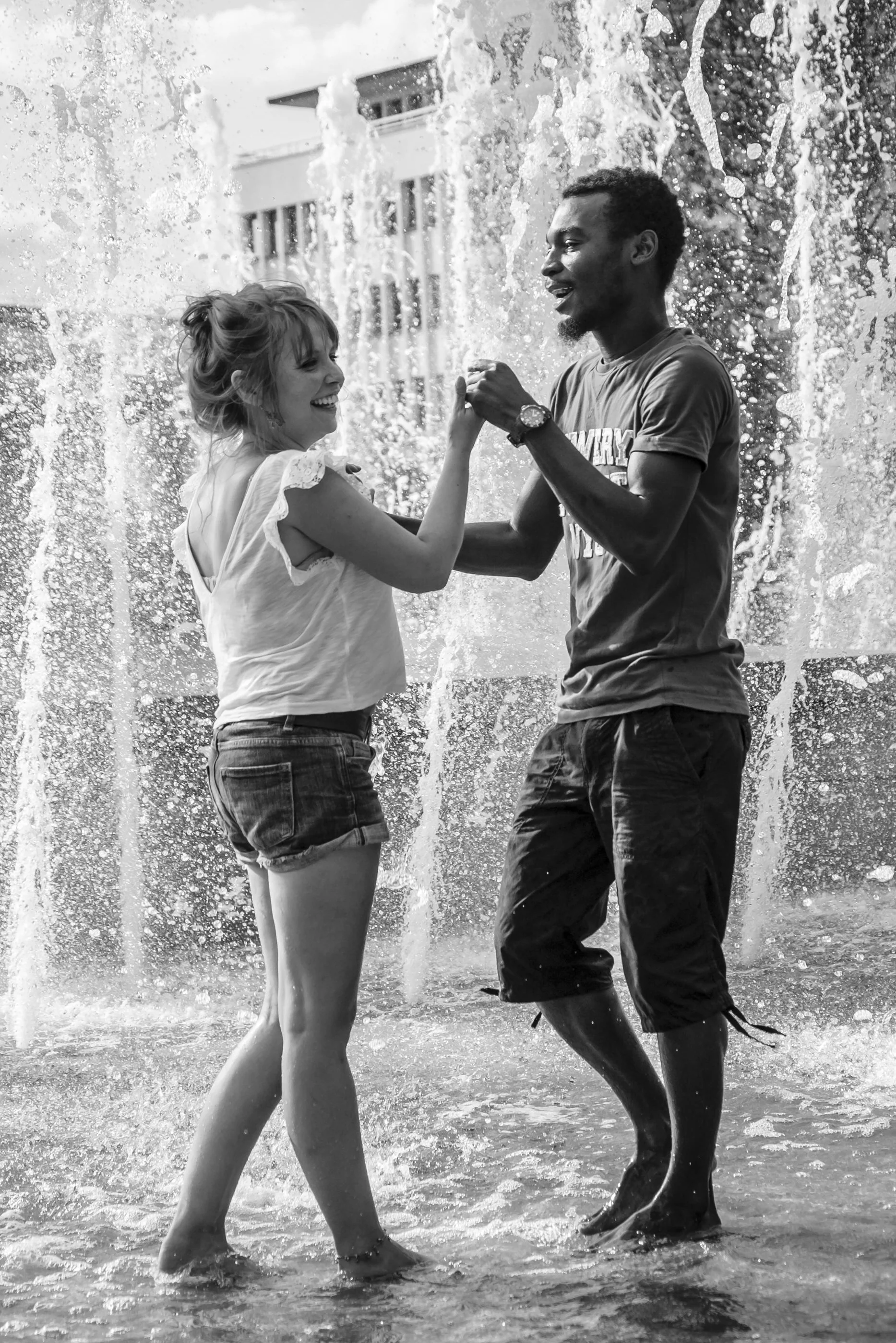 A black and white photo of a young woman and a young man dancing together in water at the beach, holding hands and smiling with water splashing around them.