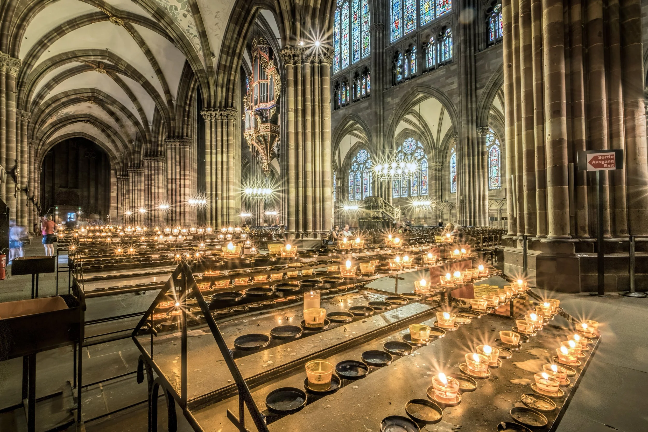 Inside a large Gothic cathedral with tall stained glass windows, stone columns, and arching ceilings. The space is illuminated by candlelight, with numerous lit candles on tables, and a small group of people is visible.