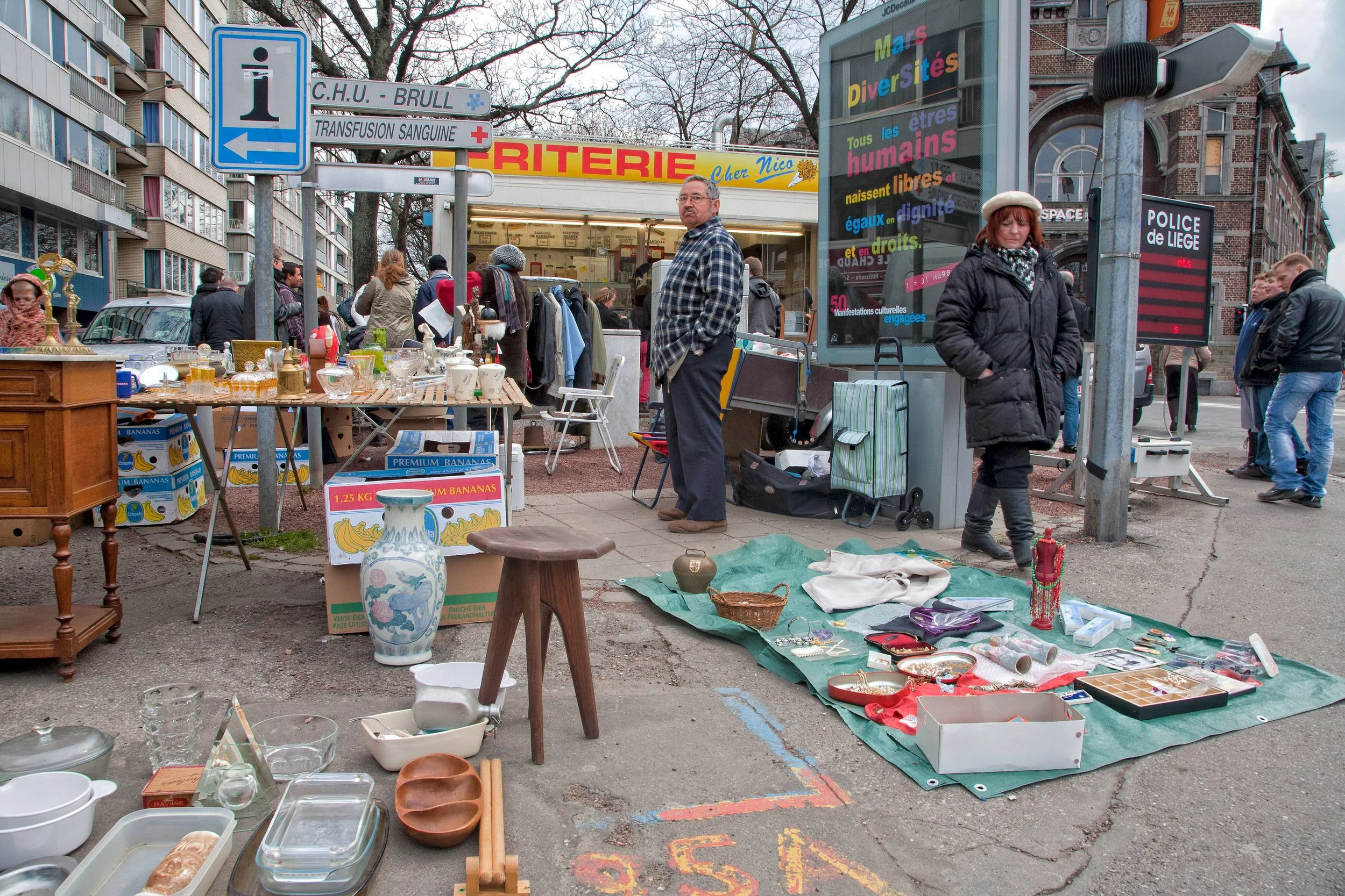 An outdoor flea market with various items on display, including ceramics, glassware, and decorative objects. People browse and shop along a street, with a sign for a charity store and buildings in the background.