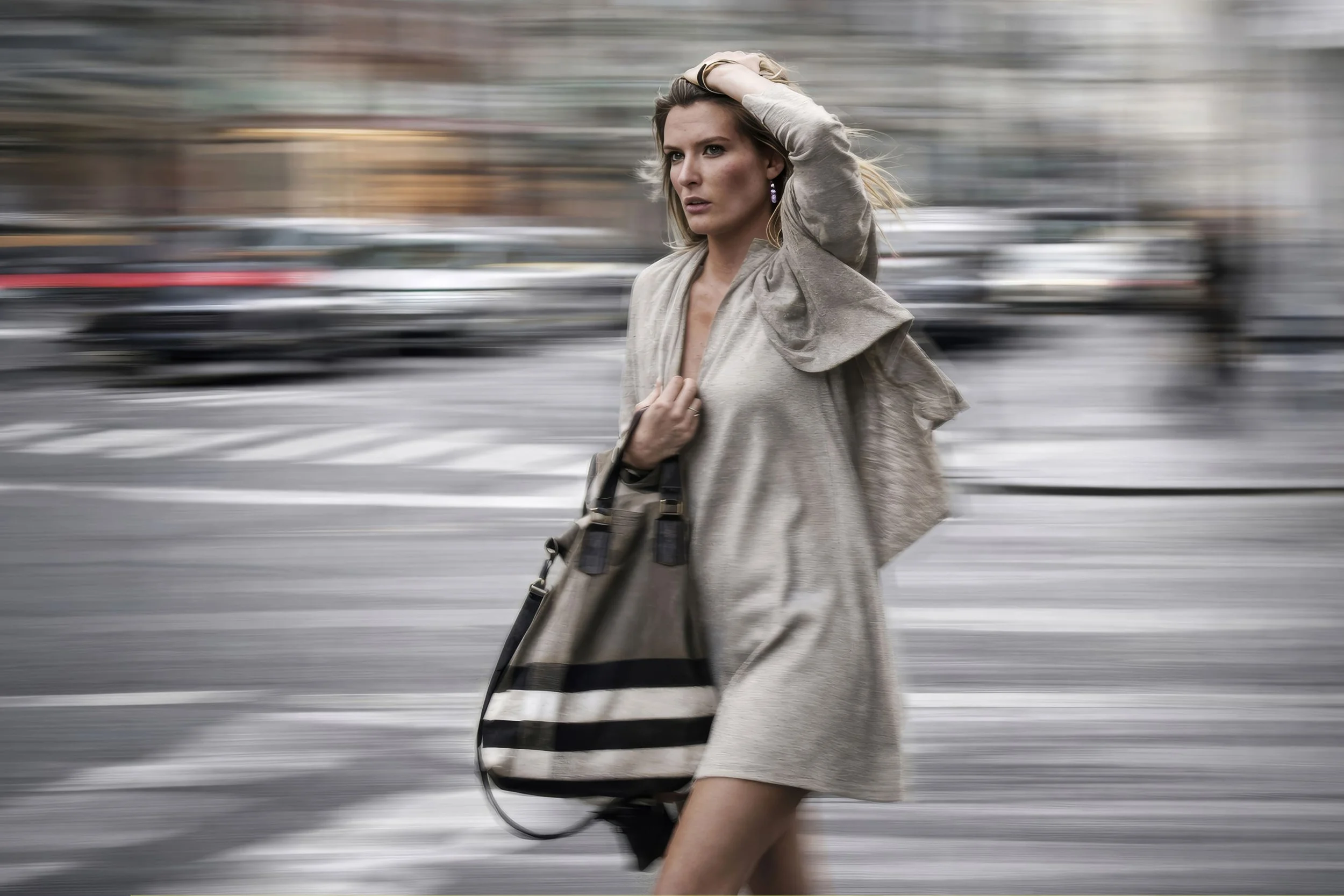 A woman walking across a busy city street with blurred cars in the background, dressed in a beige trench coat and carrying a black bag.