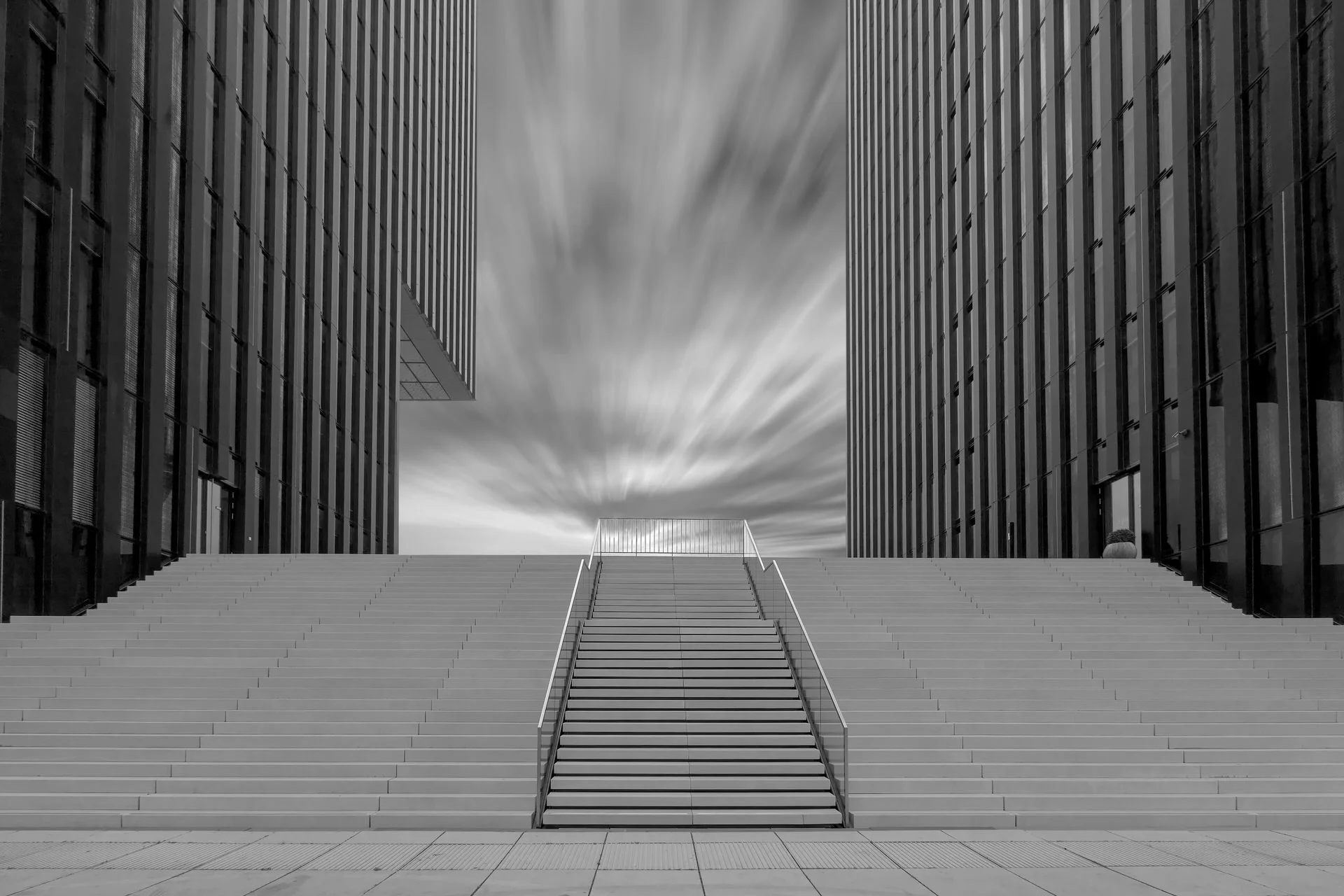 A wide black and white photograph of a modern urban scene with a staircase leading up between two towering glass and metal buildings, under a sky with long, streaked clouds.