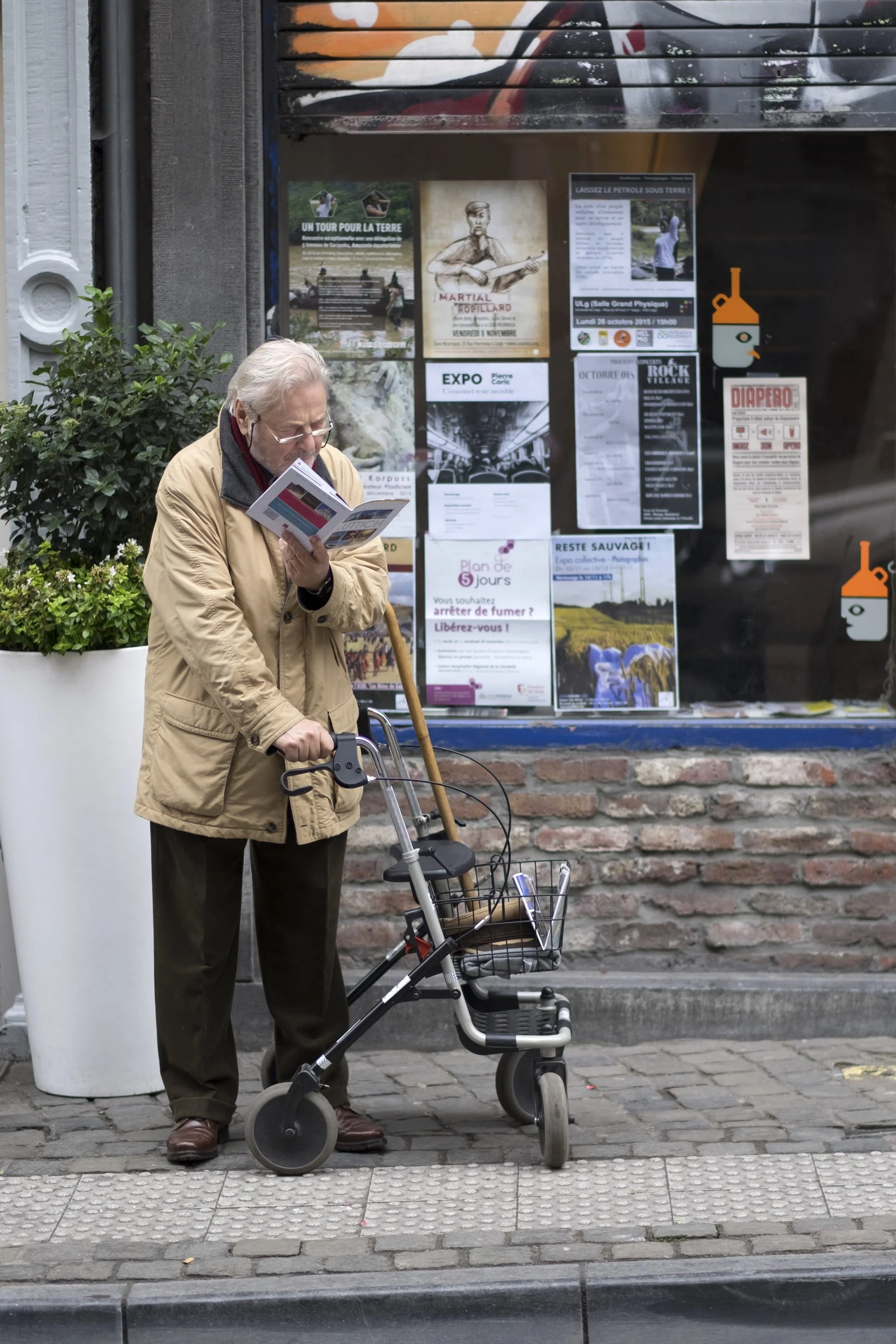 An elderly man with white hair and glasses, wearing a tan jacket, stands next to a shopping cart and uses a cane on a city sidewalk. Behind him, a large window displays various posters and flyers, with one advertising a guitar event and others promot