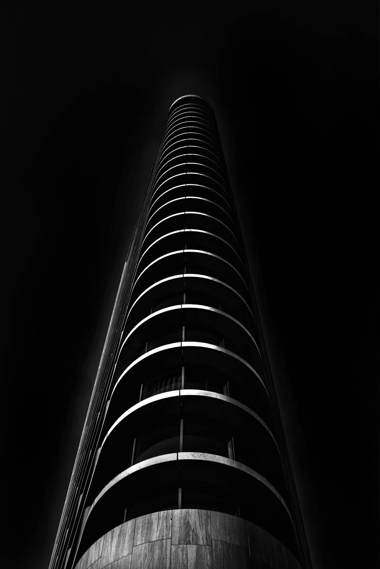 A black and white photo of a tall building with a circular architectural design, taken from the ground looking up, with a focus on the building's curved, radiating structure against a dark sky.