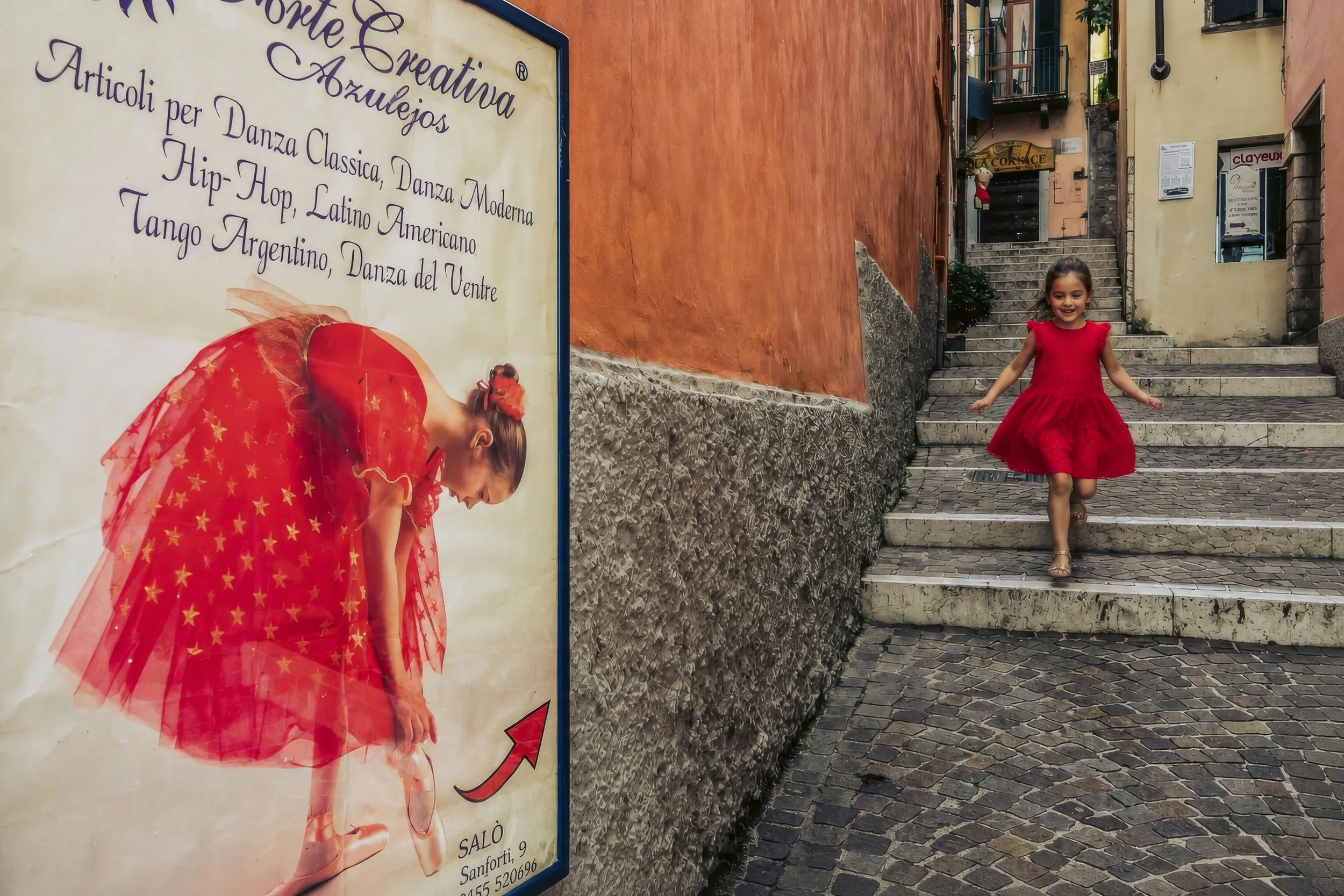 A young girl wearing a red dress and sandals walking down stone steps in a European-style street with colorful buildings. To the left, a sign with a girl in a red dress with gold stars is visible.