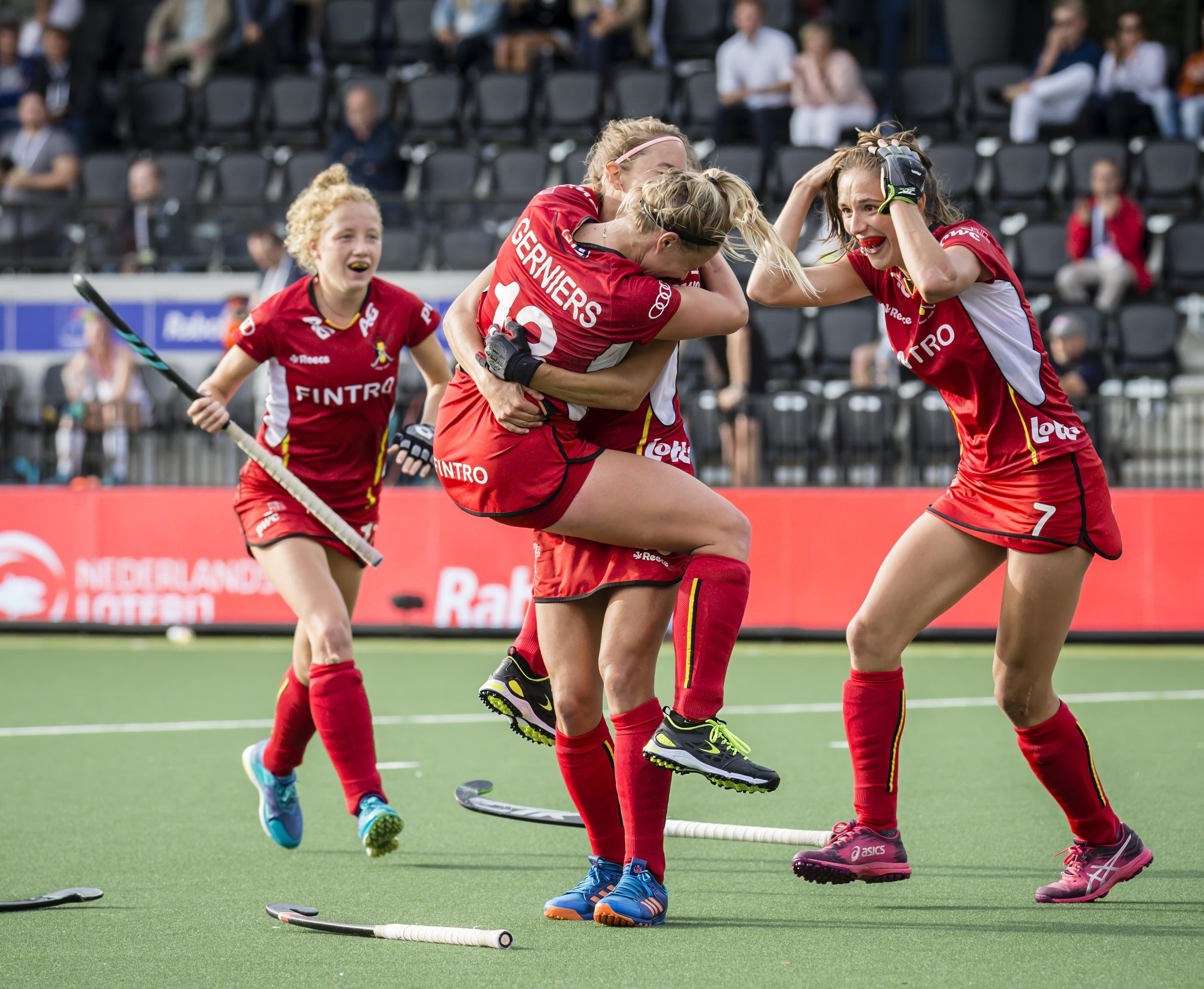 A group of female field hockey players in red uniforms celebrate on the field, with some hugging and smiling, while others stand nearby with hockey sticks.