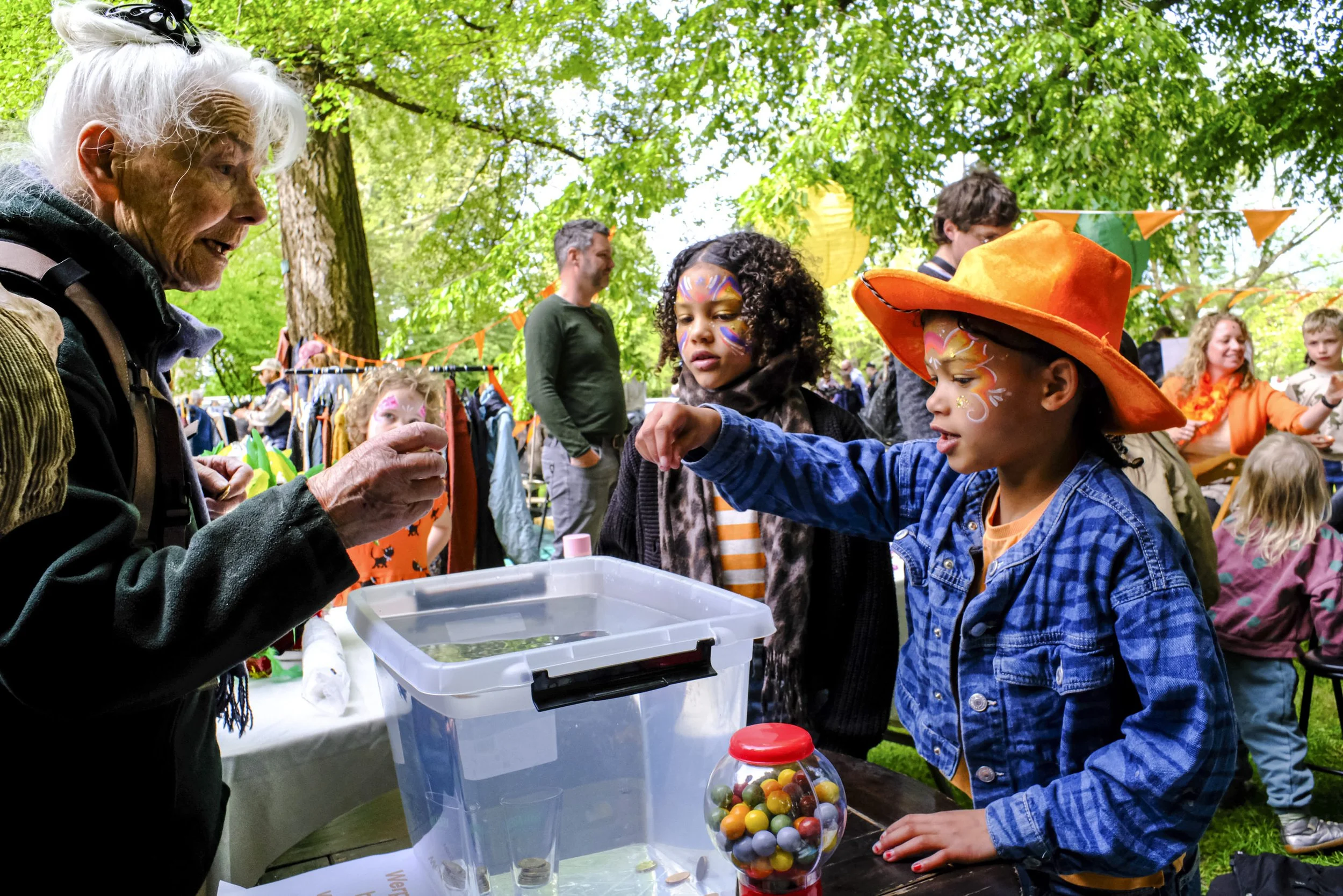 An elderly woman and a young girl with face paint at an outdoor festival, playing bingo with a transparent ball filled with colorful balls, under green trees with bunting.