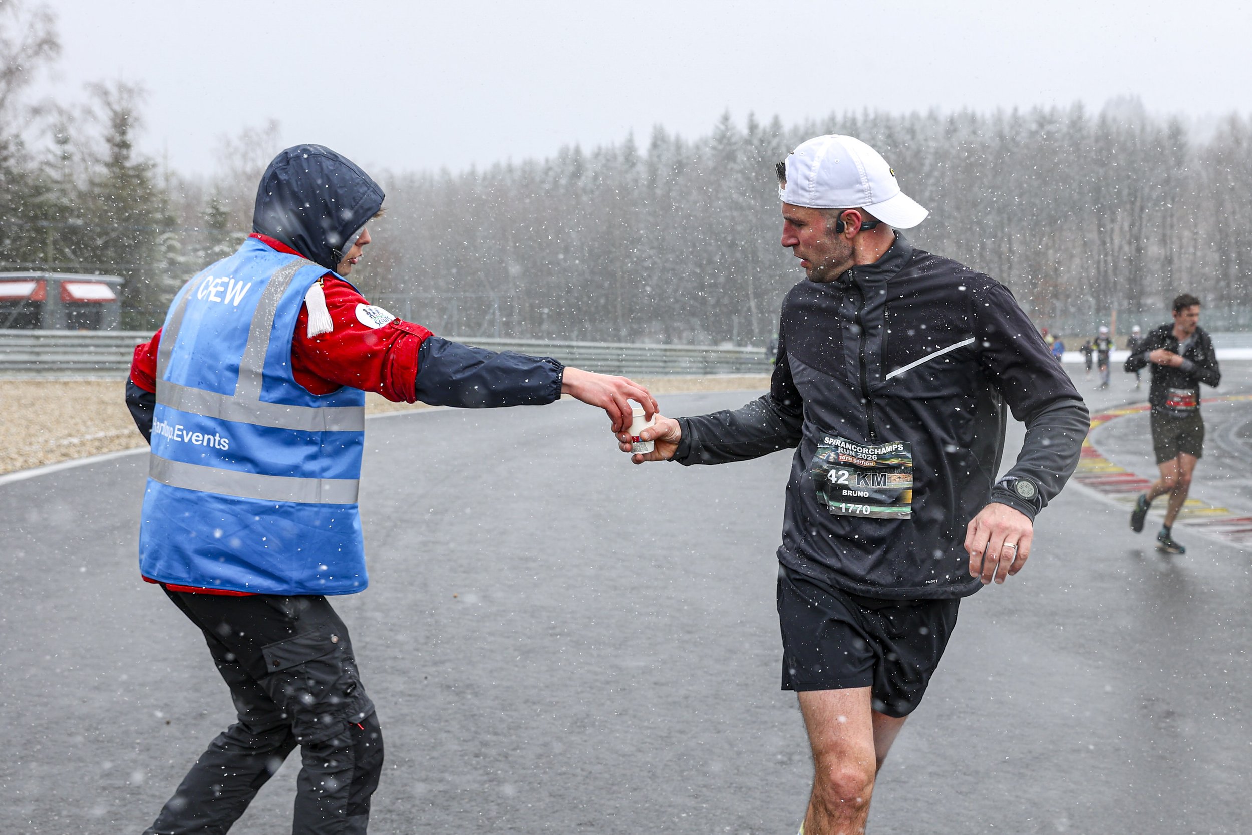 A man with a race bib and athletic gear, including a white cap, is being handed water by a race volunteer wearing a blue vest and hooded jacket in rainy weather during a race.