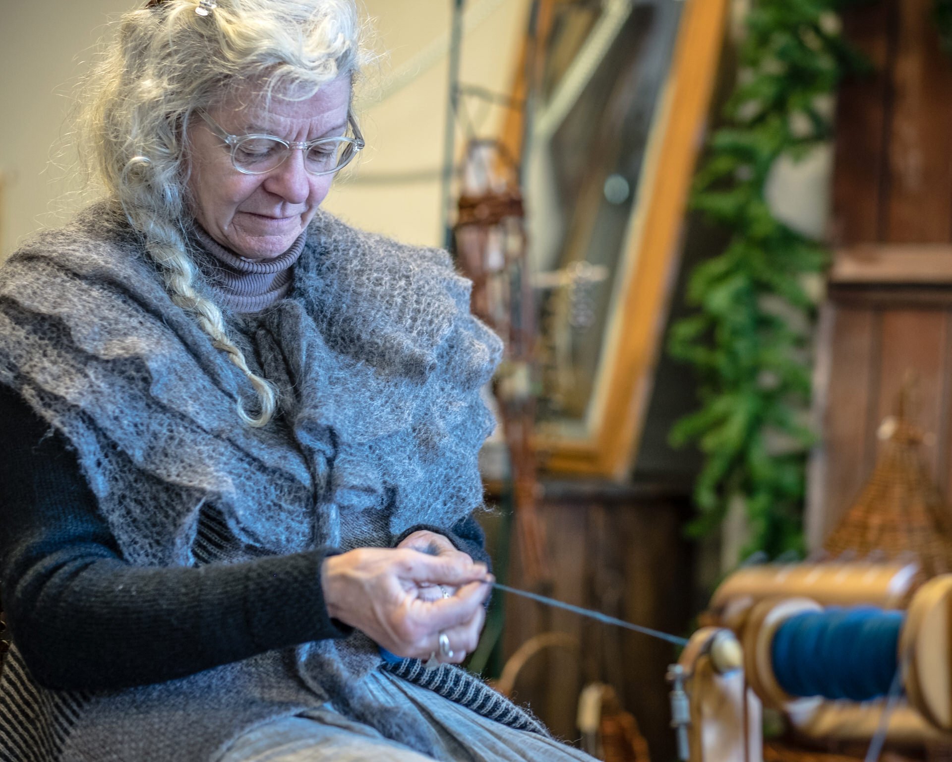 An elderly woman with curly gray hair, wearing glasses and a sweater, sitting in a cozy indoor space with plants and wooden furniture, knitting.