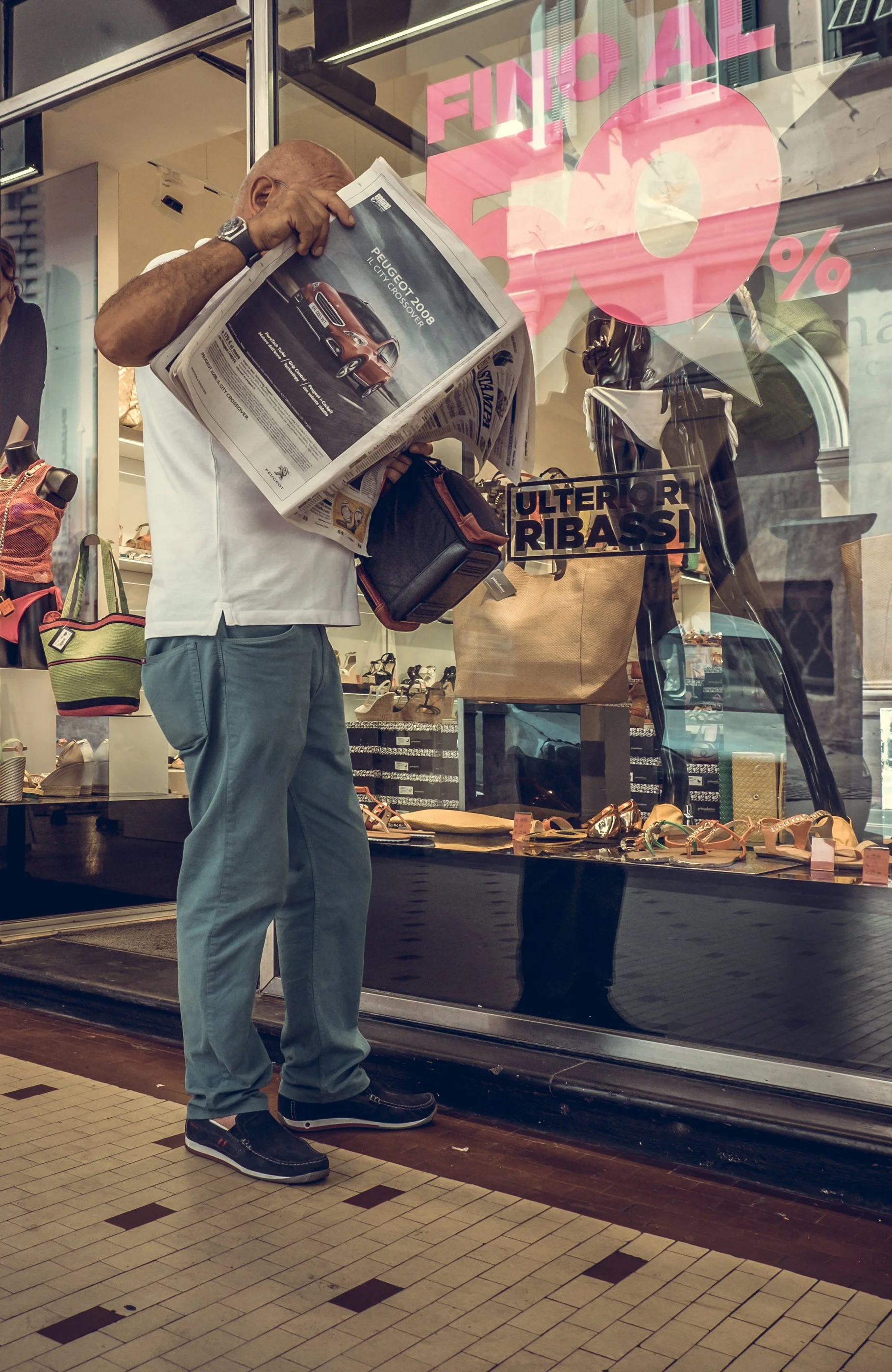 A man is standing outside a store, reading a newspaper. The store window displays a large pink sign that says 'Final 50%'. Inside the store, shelves and mannequins with shoes and handbags are visible.