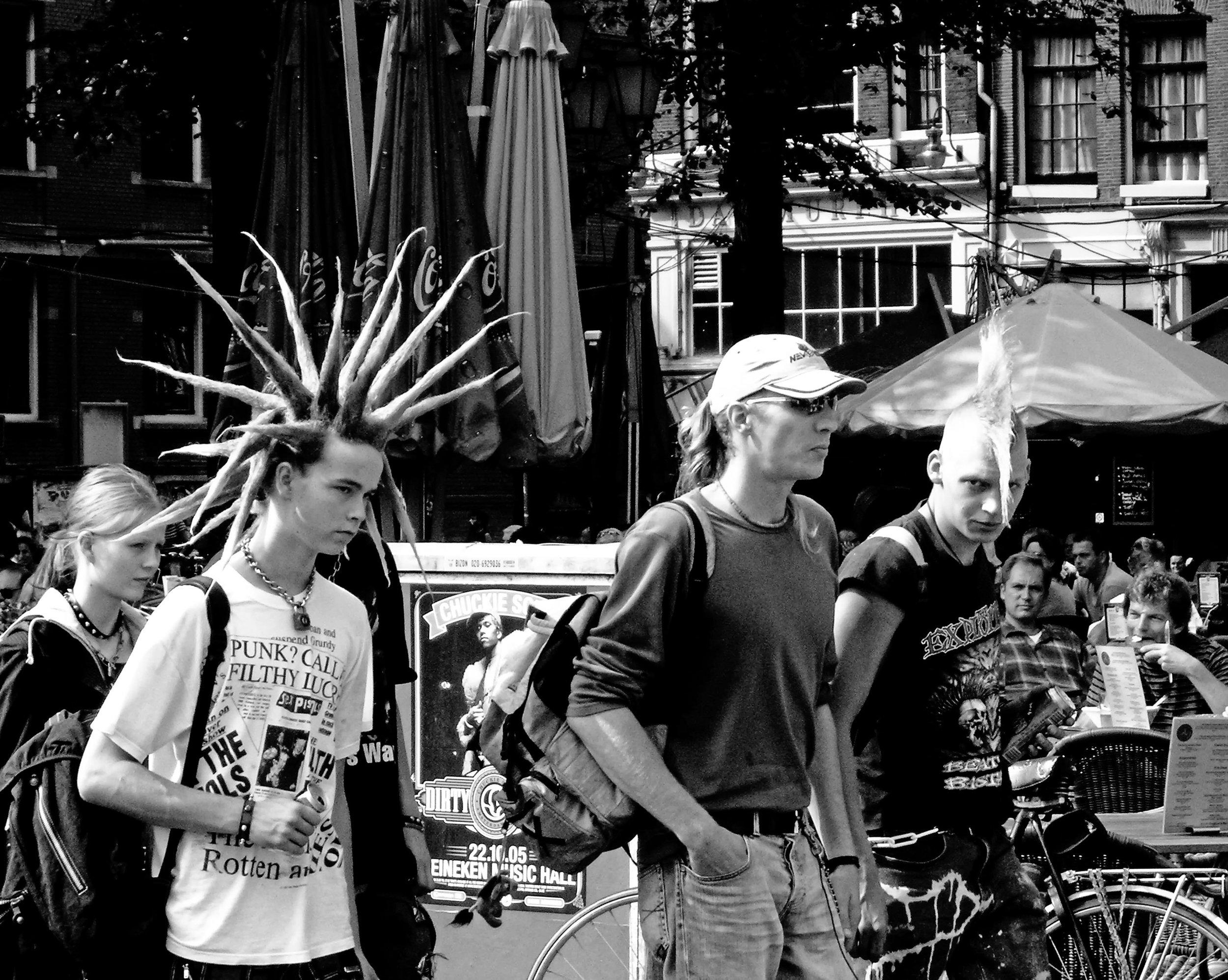 A black and white photo of people at an outdoor event, including a person with a mohawk haircut, others with various hairstyles, and a woman with sunglasses and a hat, surrounded by tents and buildings.