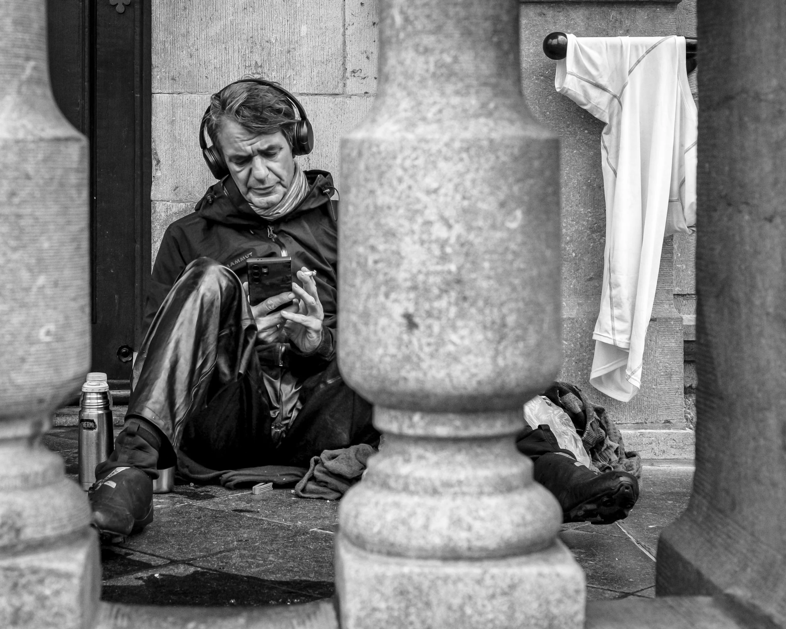 A man smoking and looking at his phone, sitting on the ground among urban stone columns, with a thermos and a bag nearby, seen through a gap between columns.