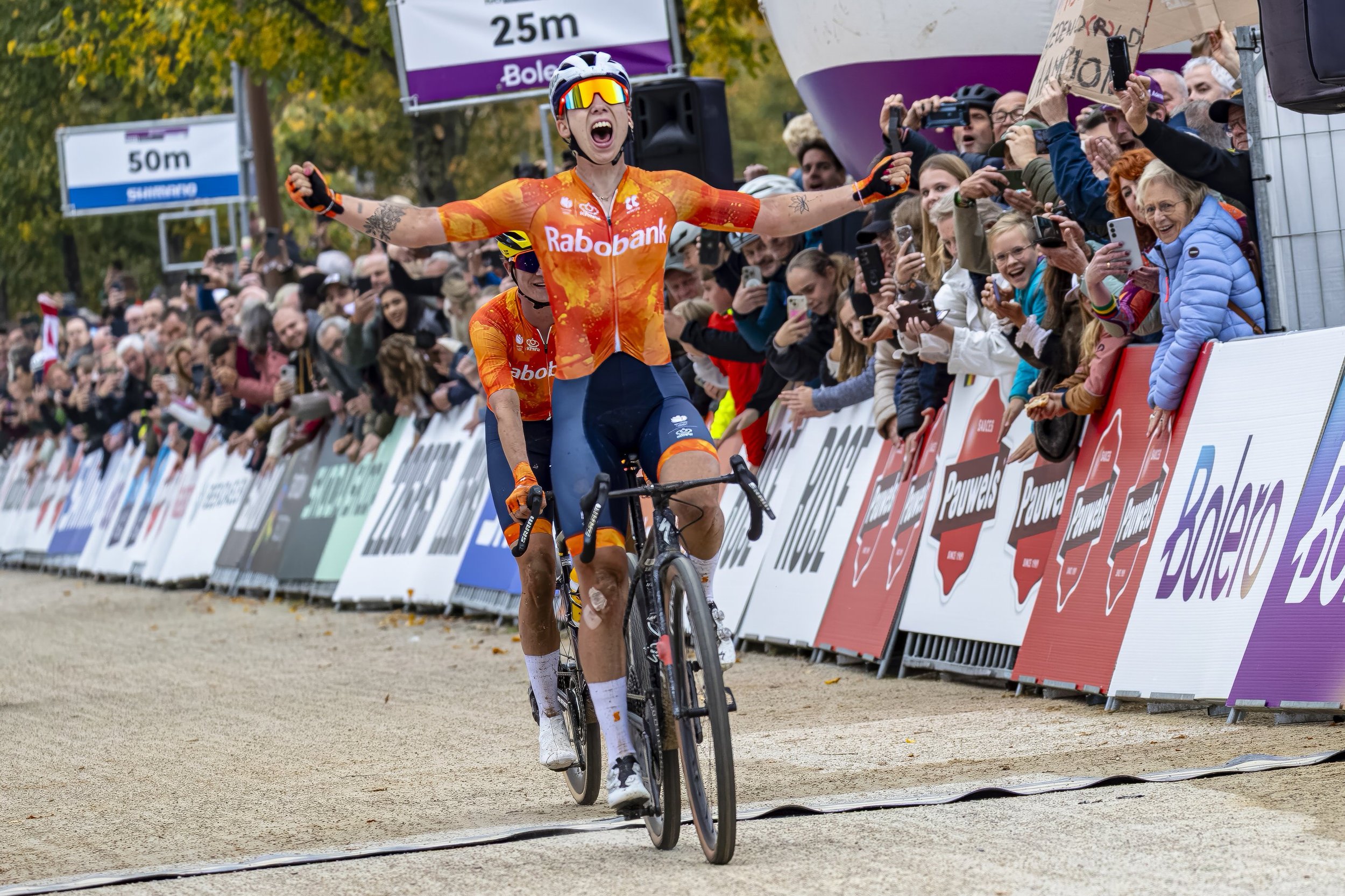 A cyclist crossing the finish line at a race, wearing a bright orange jersey with "Rabobank" written on it, with arms raised in celebration and crowd cheering on the sidelines.