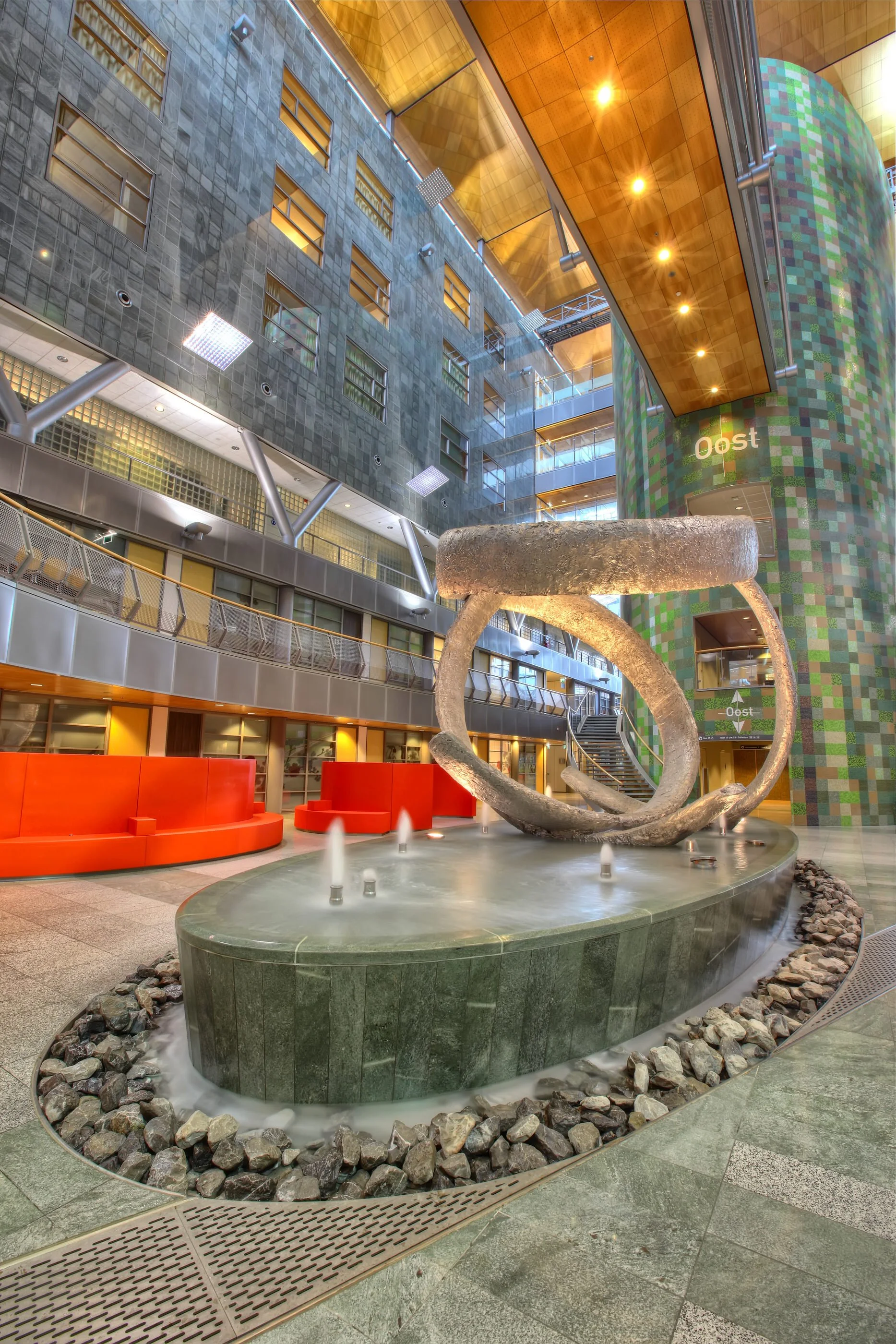 Interior of a modern building atrium with a large abstract sculpture resembling a twisted loop in the foreground, a water fountain with small jets at the base, and colorful mosaic tile walls.