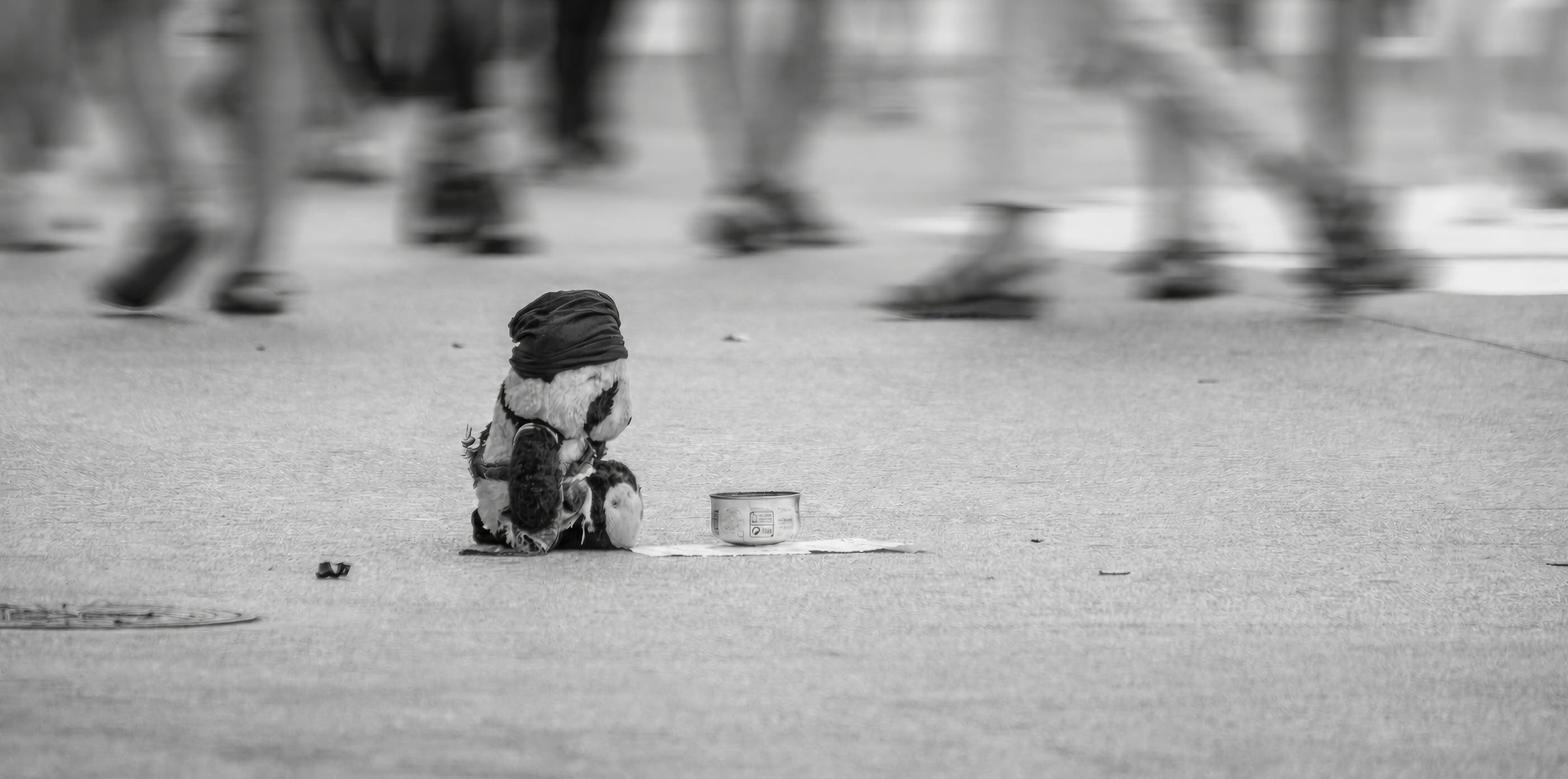 A solitary child in a hat and coat sitting on the ground near a can, with a group of blurred people walking by in the background.