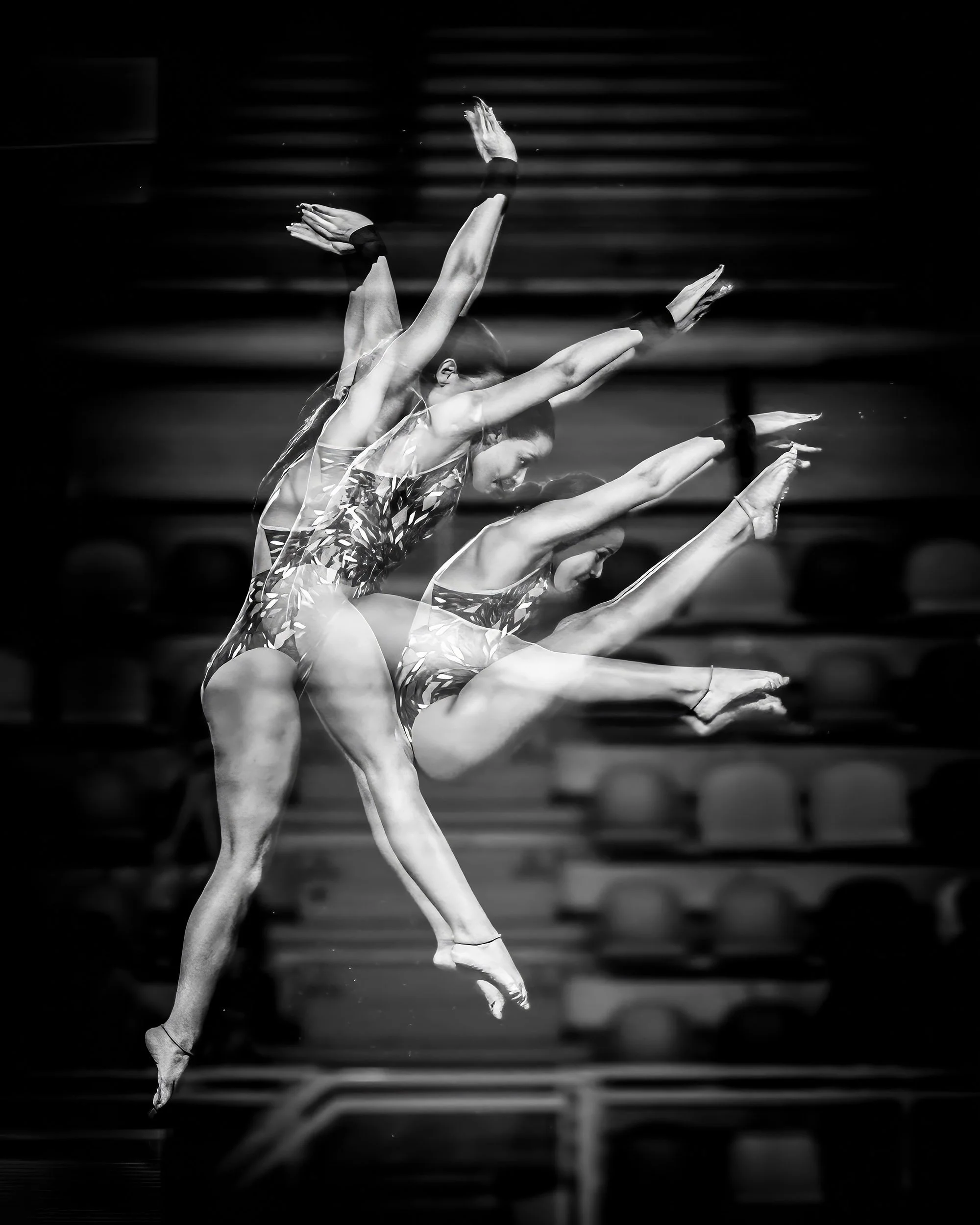 Black and white photo of a dancer mid-performance, captured with motion blur showing multiple positions of her arms and legs in a dance pose, with steps in the background.