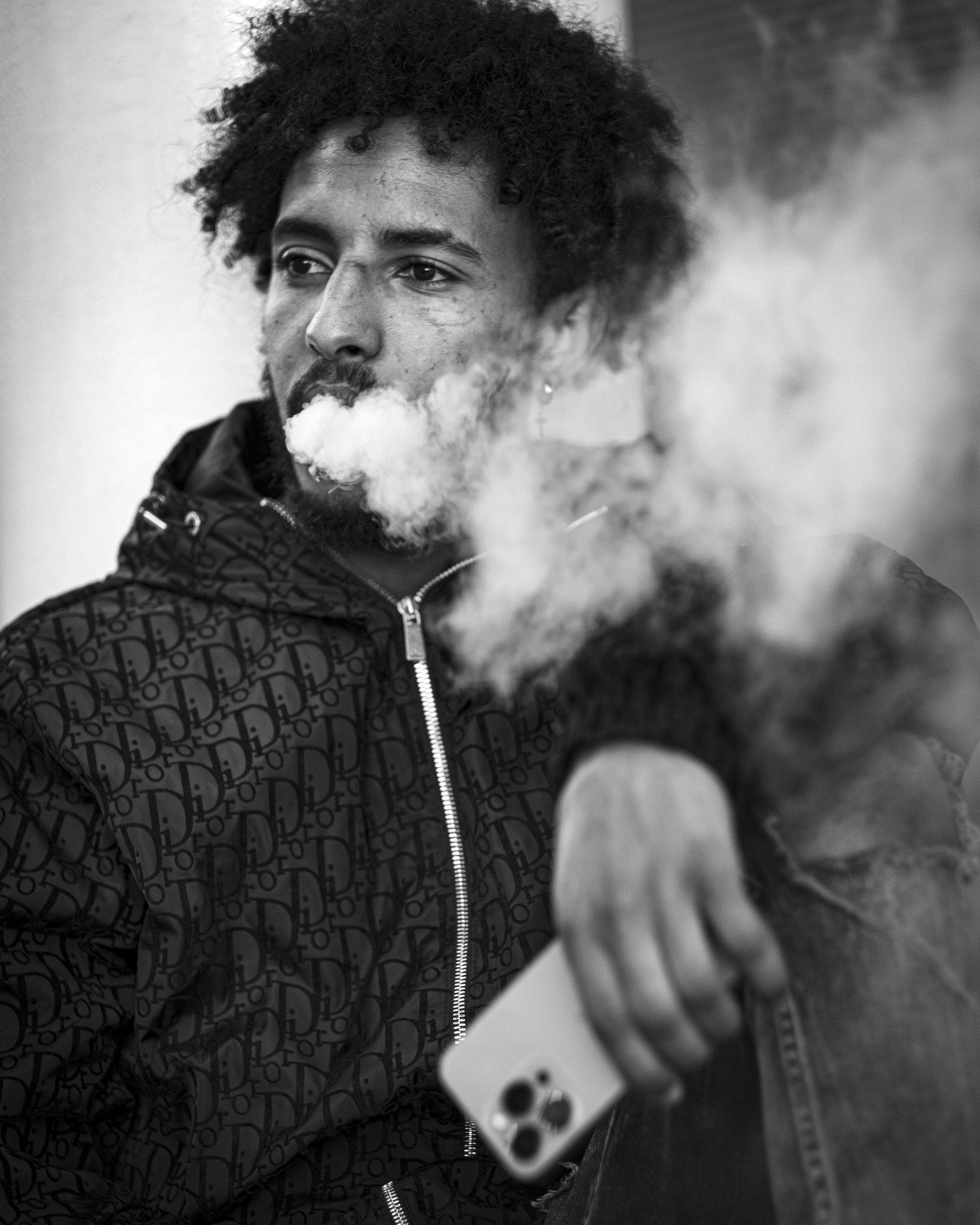 Black and white photo of a man with curly hair smoking, wearing a patterned jacket and holding a smartphone, with a skateboard in the foreground.