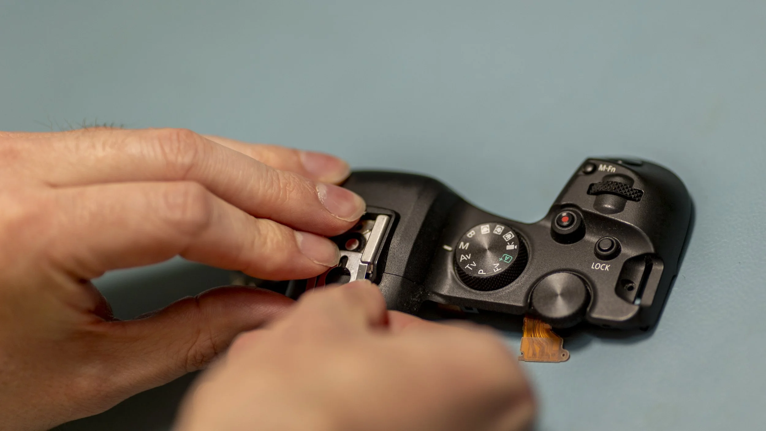A person is holding a black digital camera and appears to be changing the battery or memory card compartment, with the camera placed on a flat surface against a light blue background.