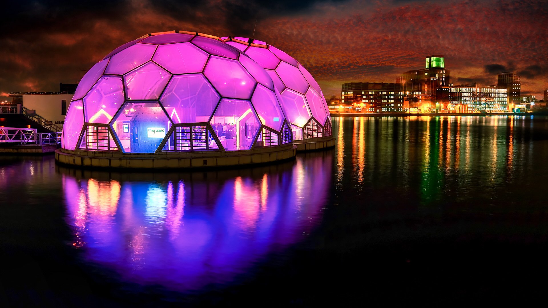 A large, illuminated geodesic dome with purple and pink lights, floating on water at night, with city buildings and colorful lights reflecting on the water in the background.