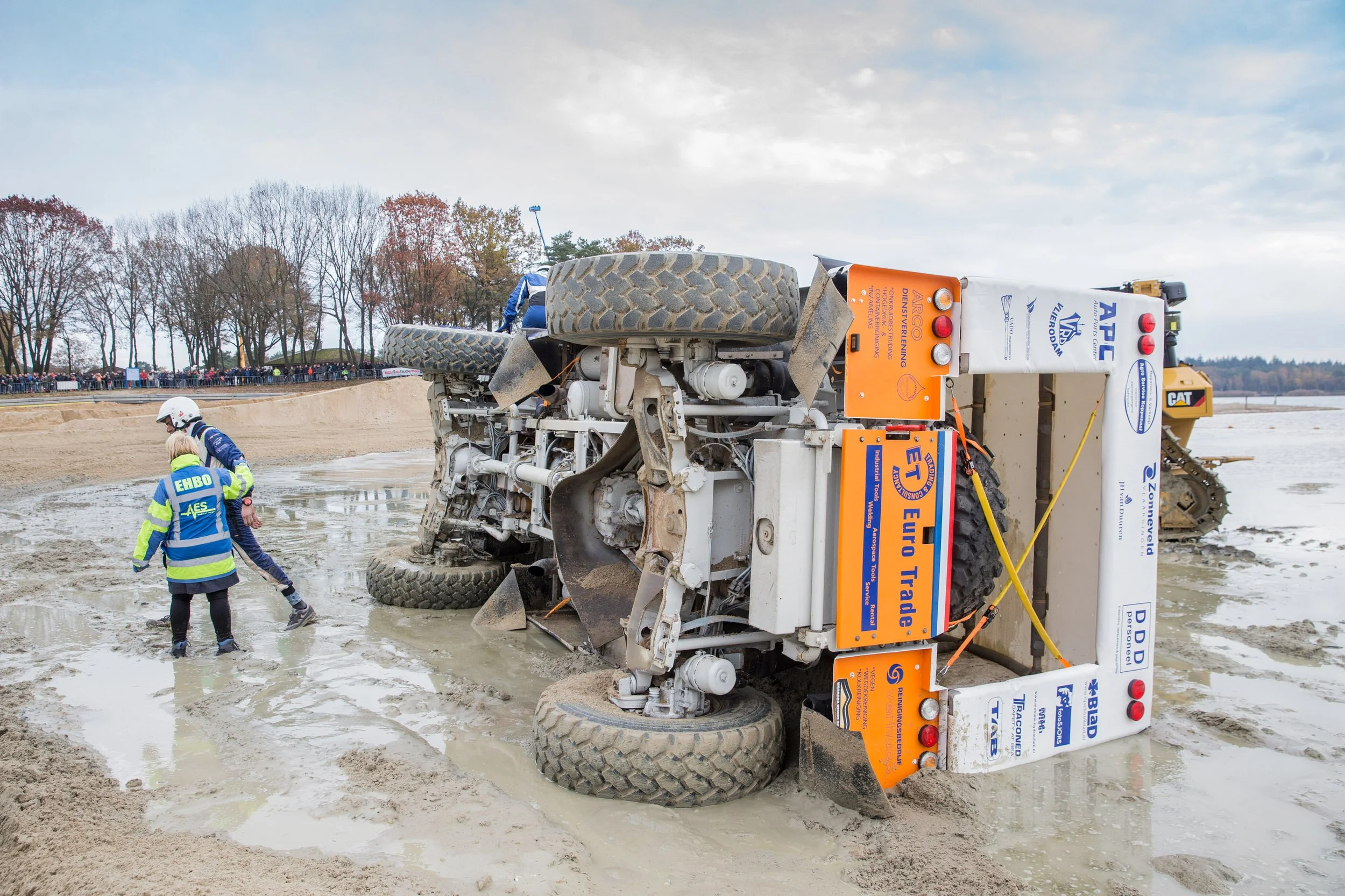 A large construction vehicle tipped over onto its side in a muddy area, with people and a crowd observing nearby.