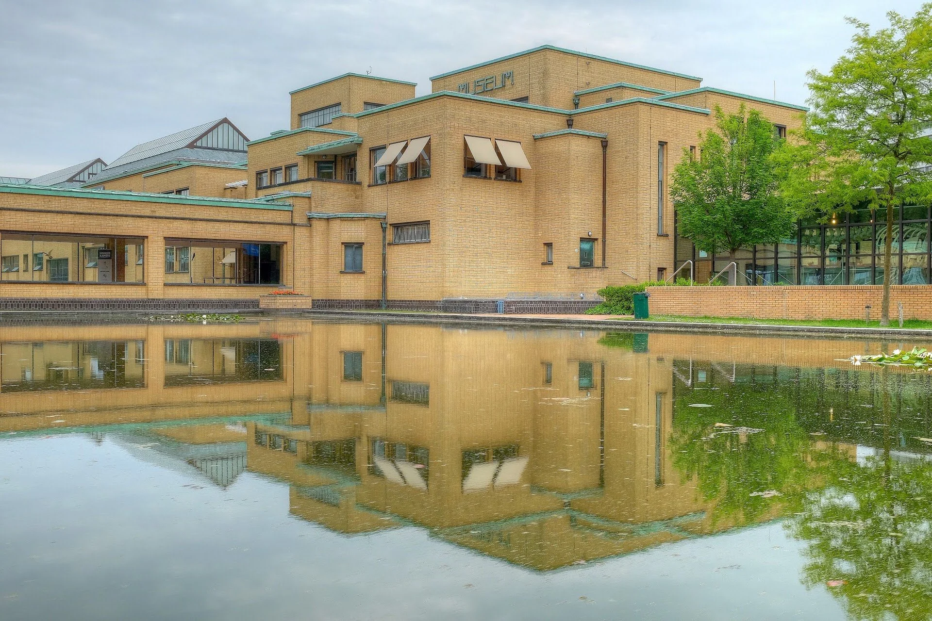 A large yellow brick museum building by a body of water, with multiple levels and cantilevered sections, green roof accents, and large glass windows reflecting the building and trees, with a tree on the right side.