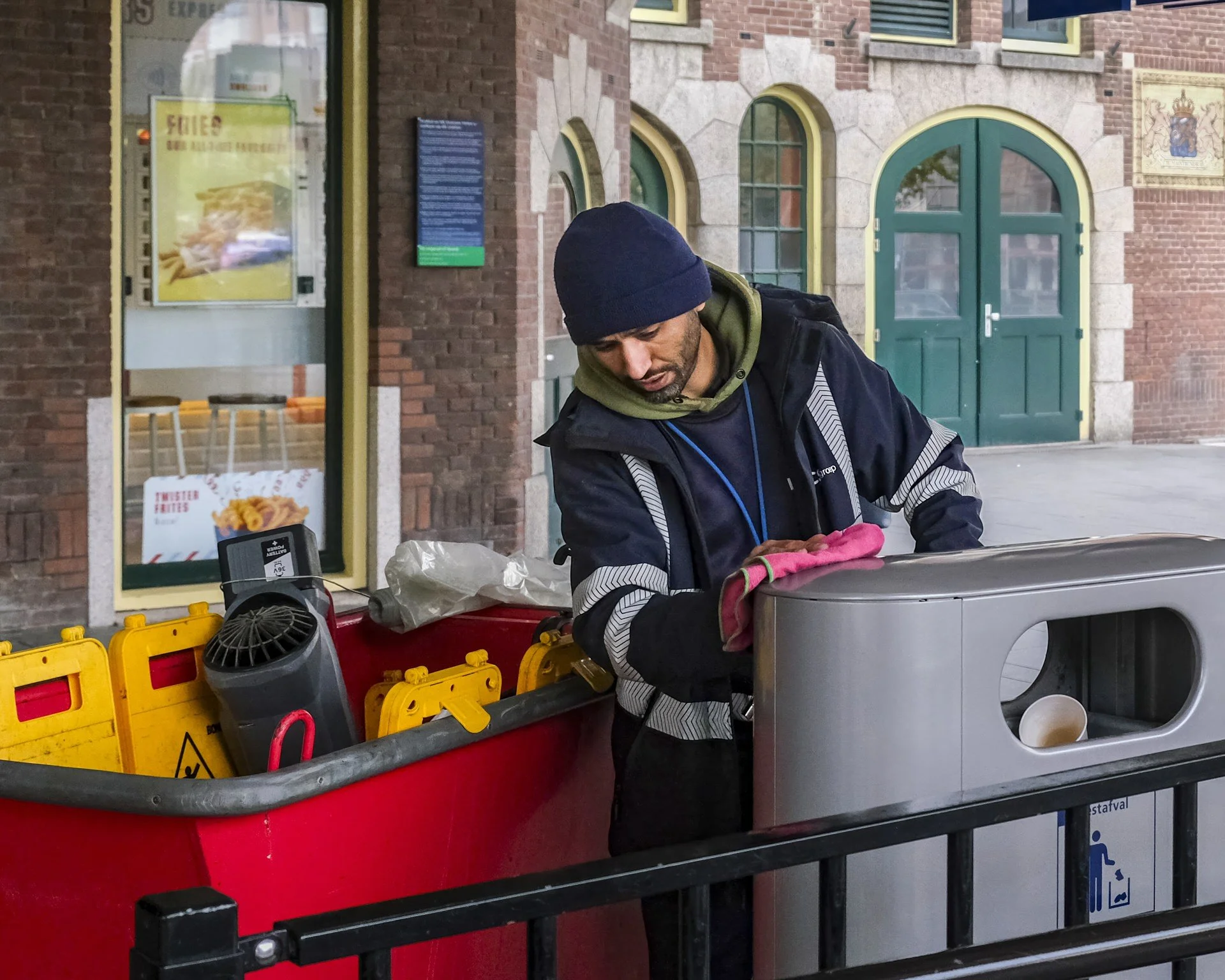 A man with a navy beanie and dark jacket working on a street repair station with various tools, inside a city with brick buildings and a green door in the background.