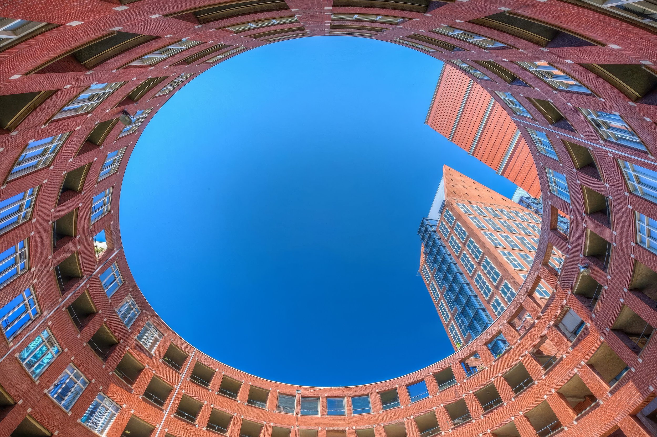 View looking up at red brick residential building with curved shape and multiple windows, against a clear blue sky.