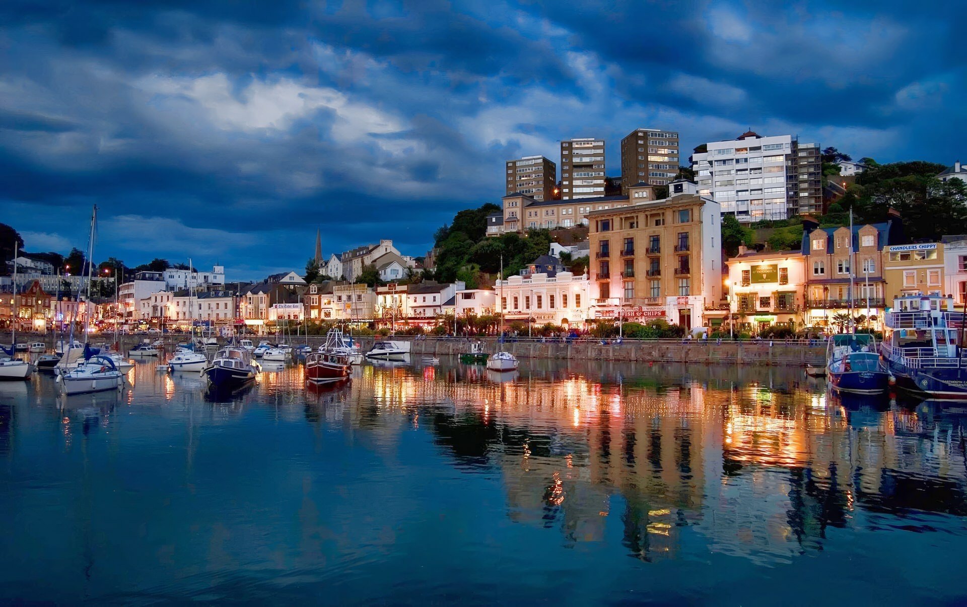 Harbor with boats and sailboats docked, buildings and homes on a hillside, and a cloudy evening sky reflected in the water.