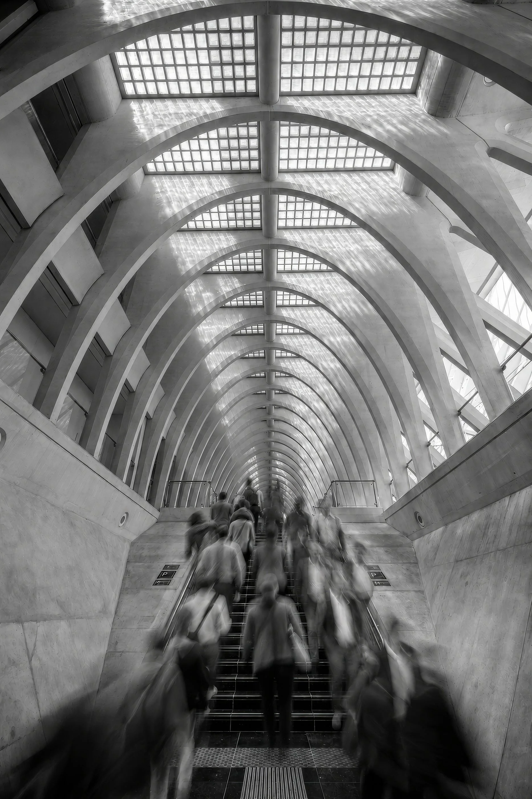 People descending an escalator in a modern, architectural underground walkway with curved concrete arches and large glass block windows.