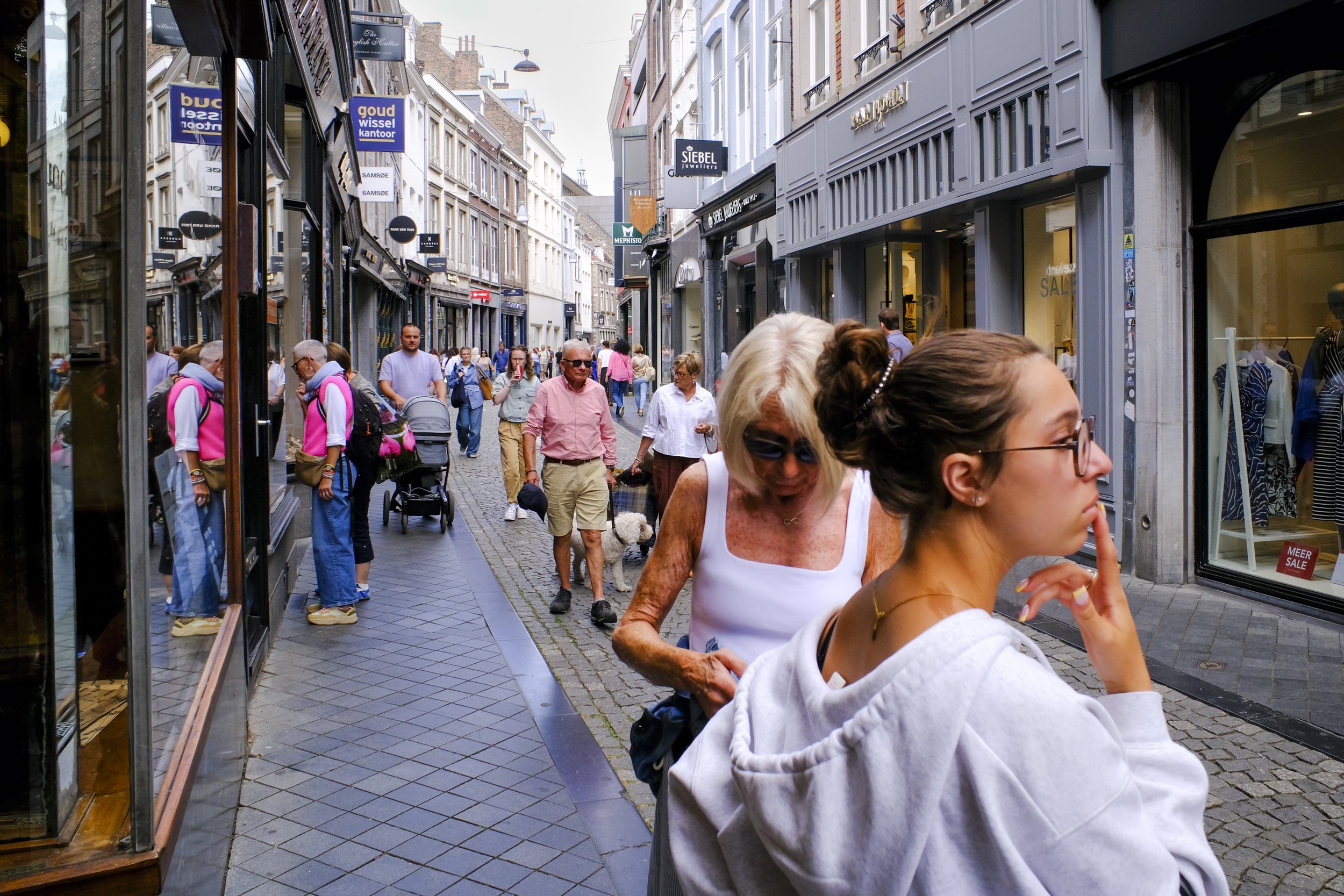 A busy street scene in a shopping district with people walking, some pushing strollers, and storefronts with signs, including a jewelry store and a sale sign.