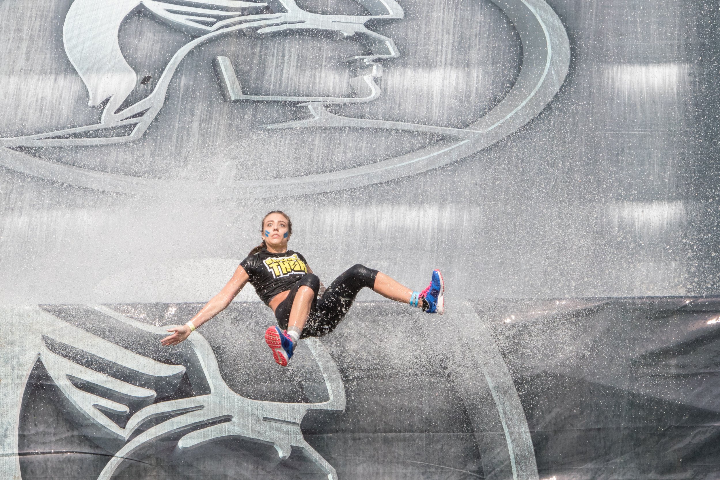 A woman in athletic clothes falling on water with splashes, on a wet surface with a large logo or design in the background.