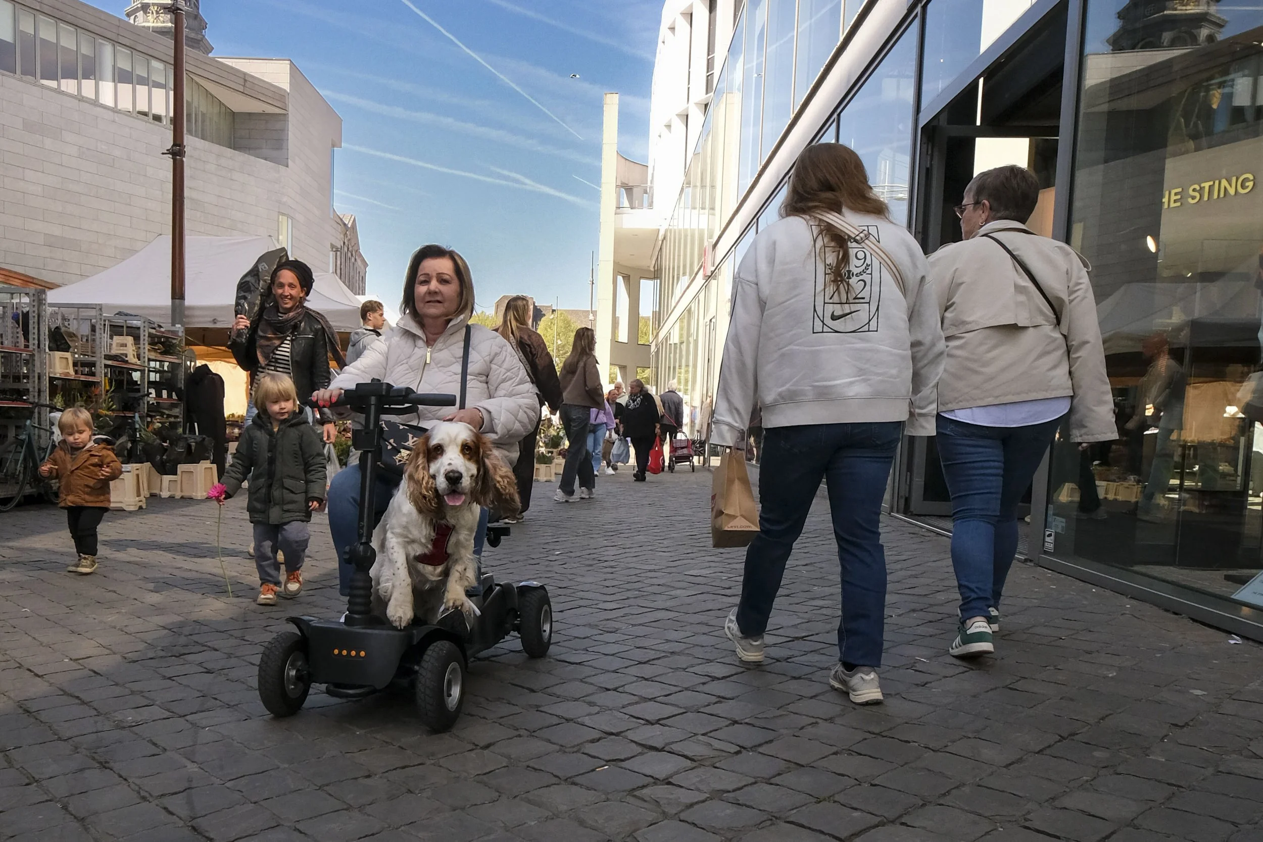 People walking on a cobblestone street in an urban area during the day. In the foreground, a woman is sitting on a scooter with a dog in front of her and two children walking nearby. Other pedestrians are visible in the background. Modern buildings a