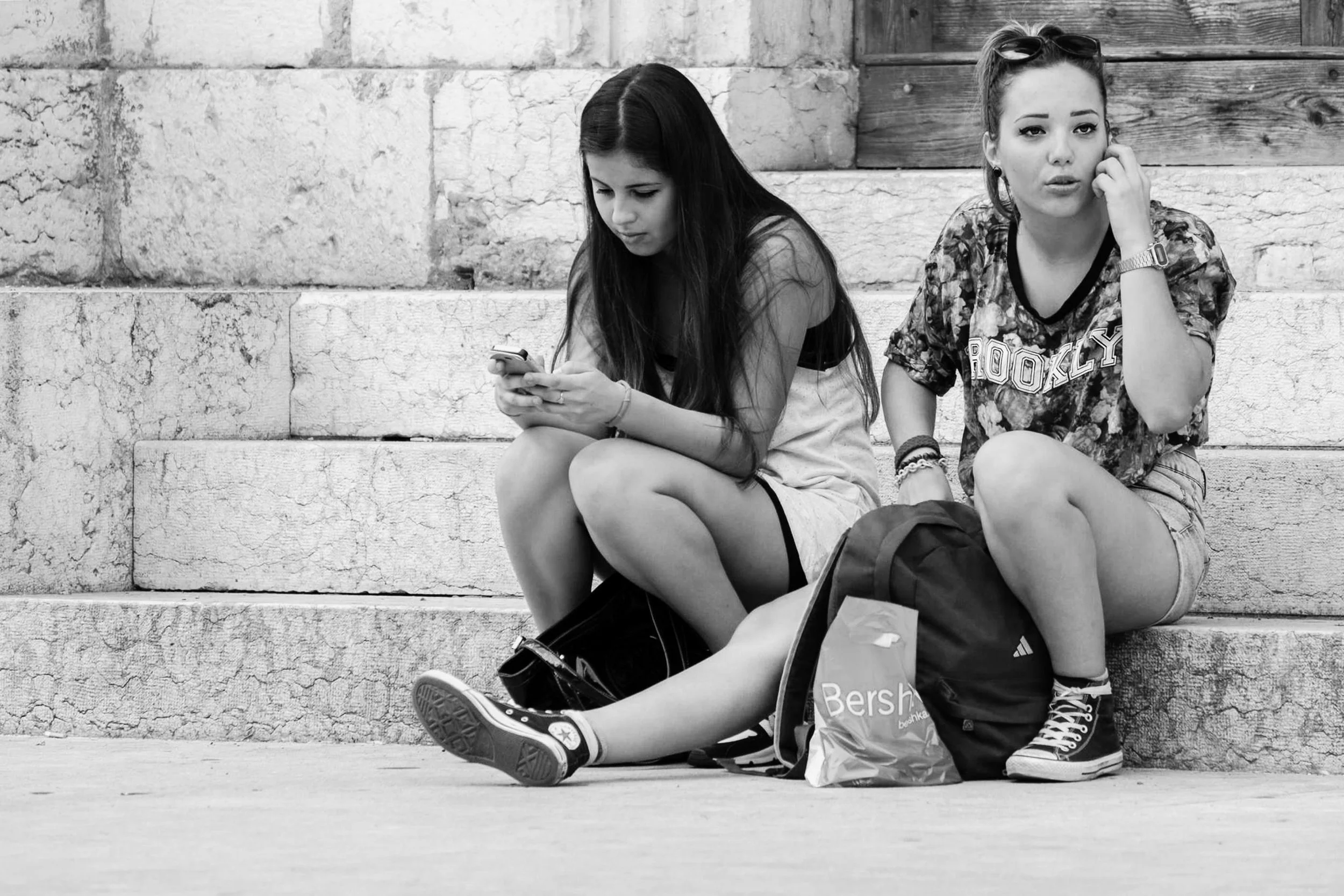 Two young women sitting on steps made of stone and wood, one looking at her phone, the other looking at the camera, with bags and belongings beside them.