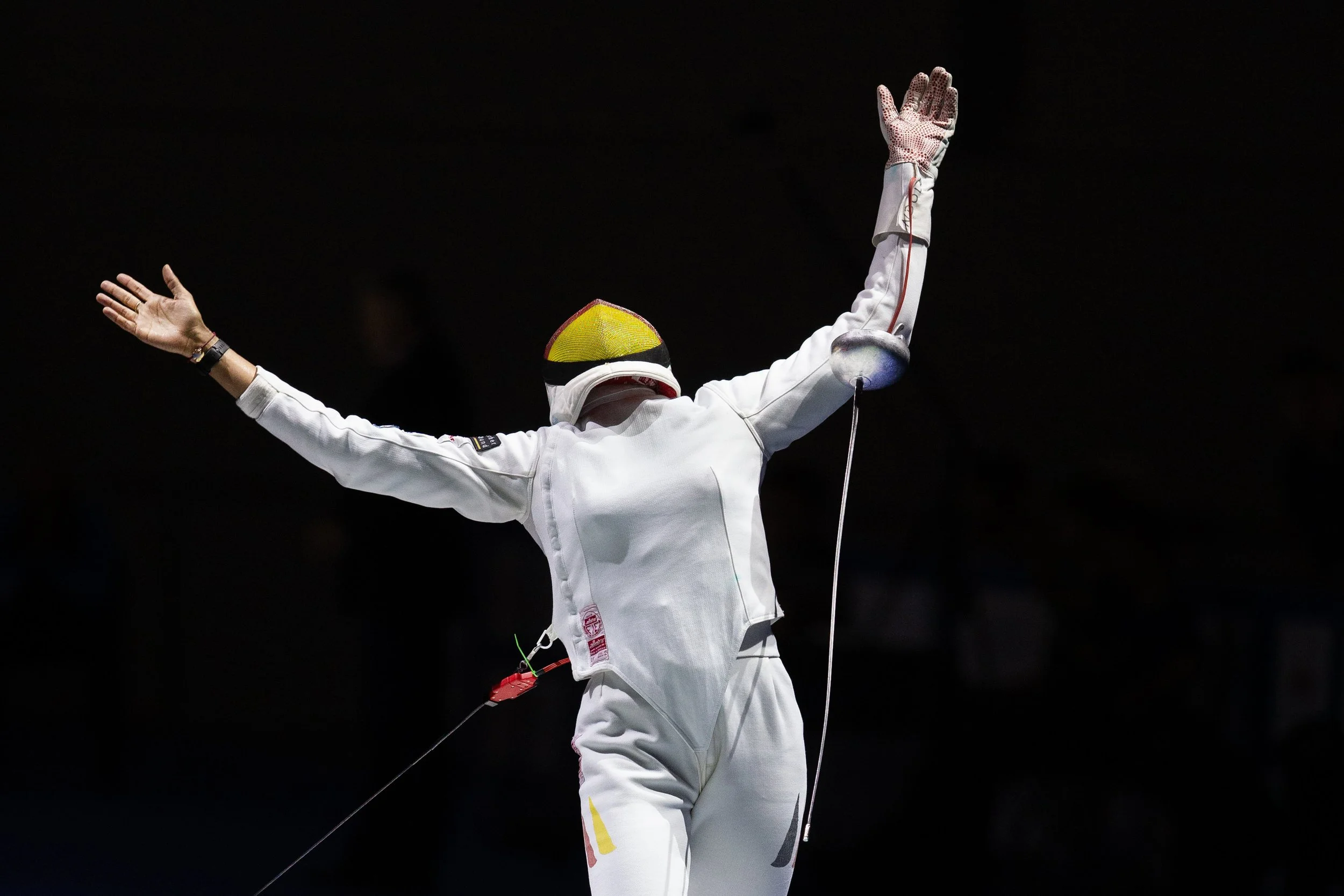 Fencer celebrating with arms outstretched and a raised fist in a fencing uniform and mask, against a dark background.