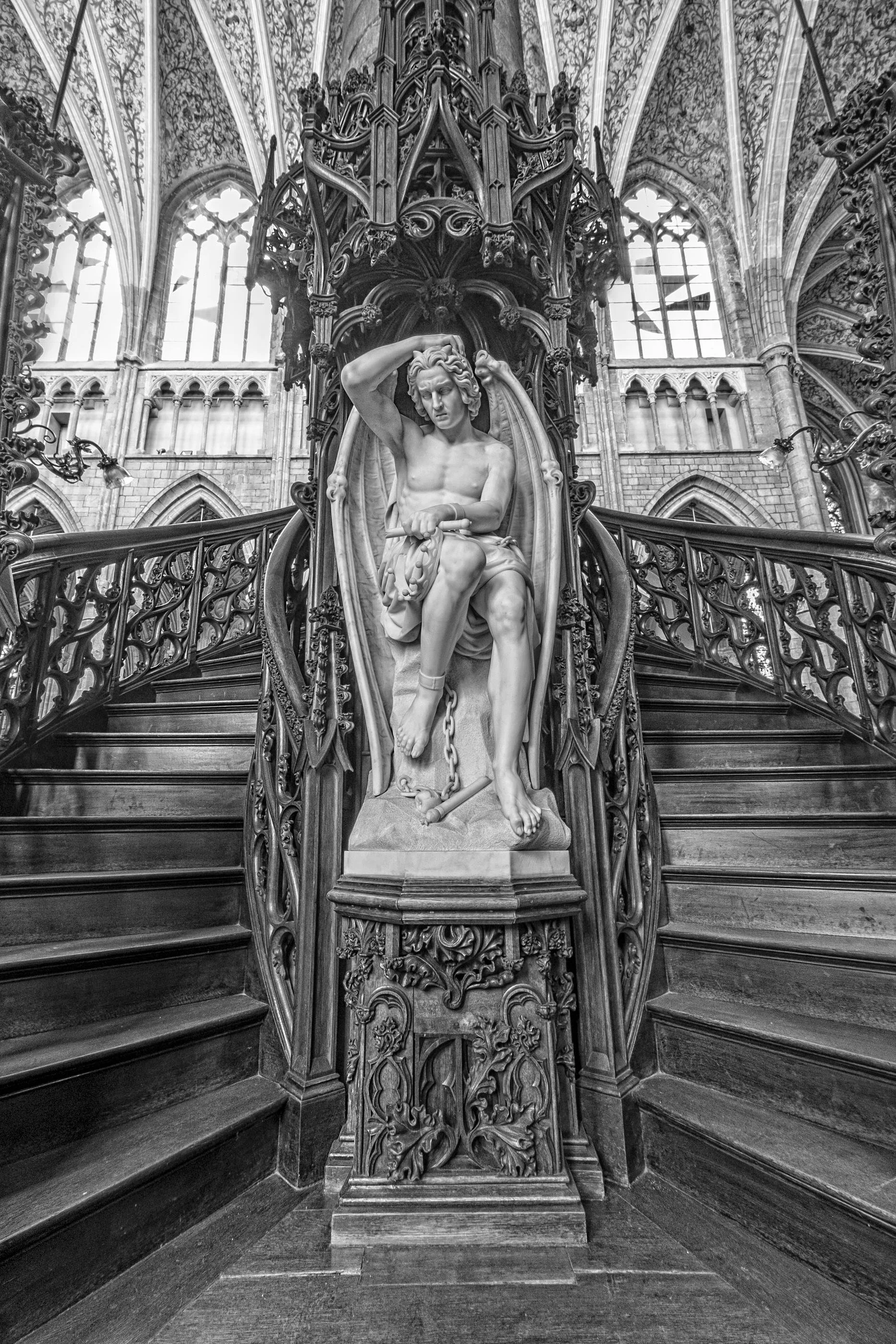 Black and white photo of a detailed Gothic interior with large stained glass windows, a grand staircase, and a marble statue of a seated man with a chain at his feet.