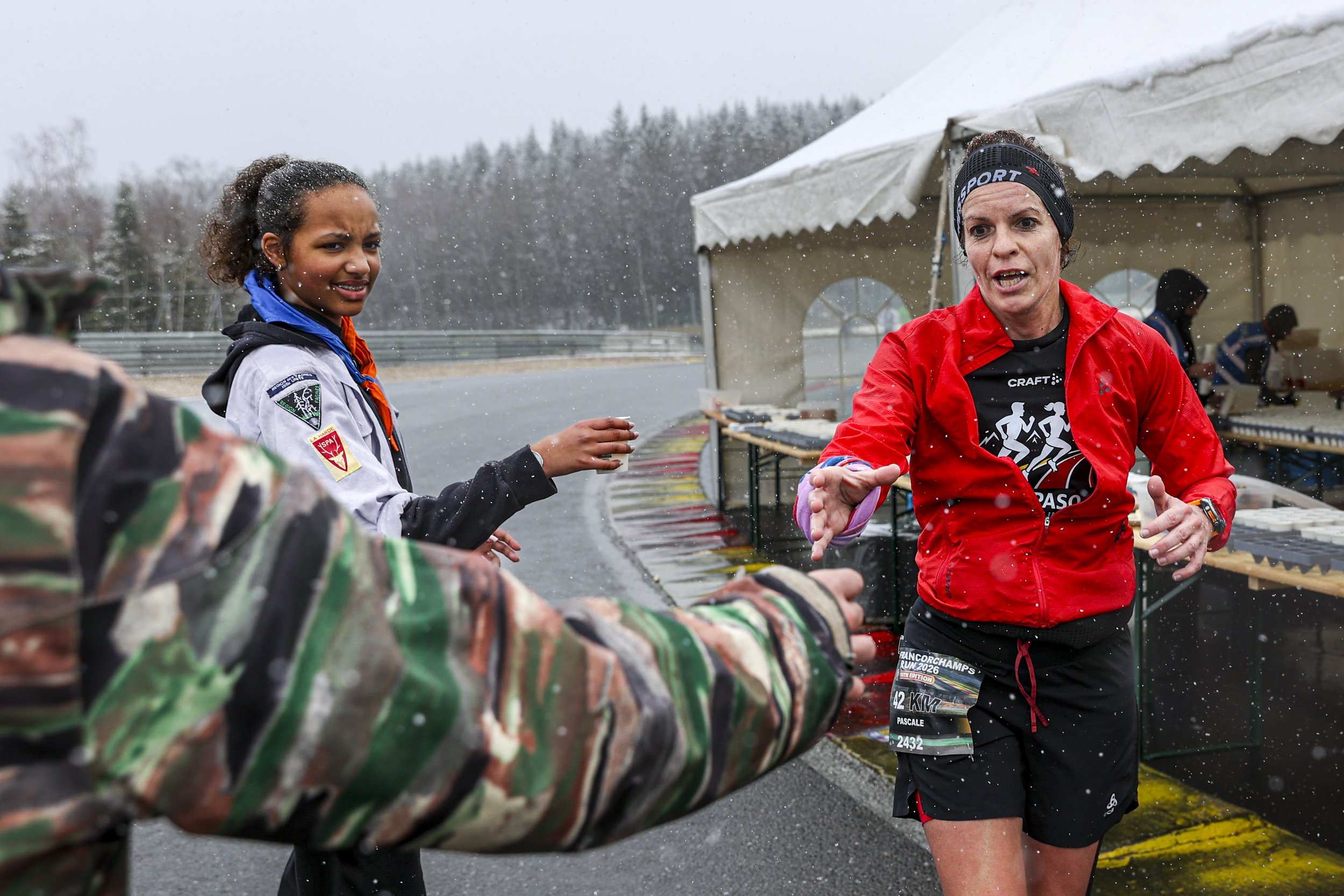 Two women participating in an outdoor race in snowy weather, reaching out to shake hands. One woman is wearing a red jacket and black shorts, the other in a white jacket. There is a tent in the background and snow on the ground.
