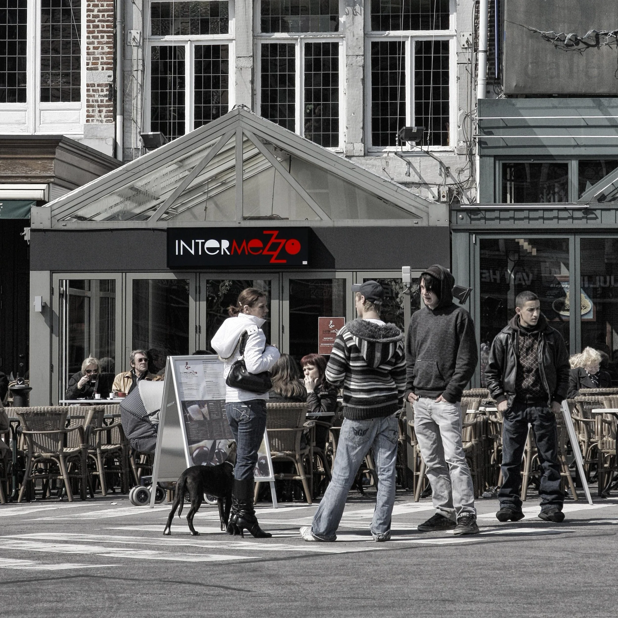 People walking on a city street outside a store with a sign reading 'INTERMEZZO' in red and white letters. A woman with a dog is in the foreground, and a group of people in casual clothing are gathered nearby.