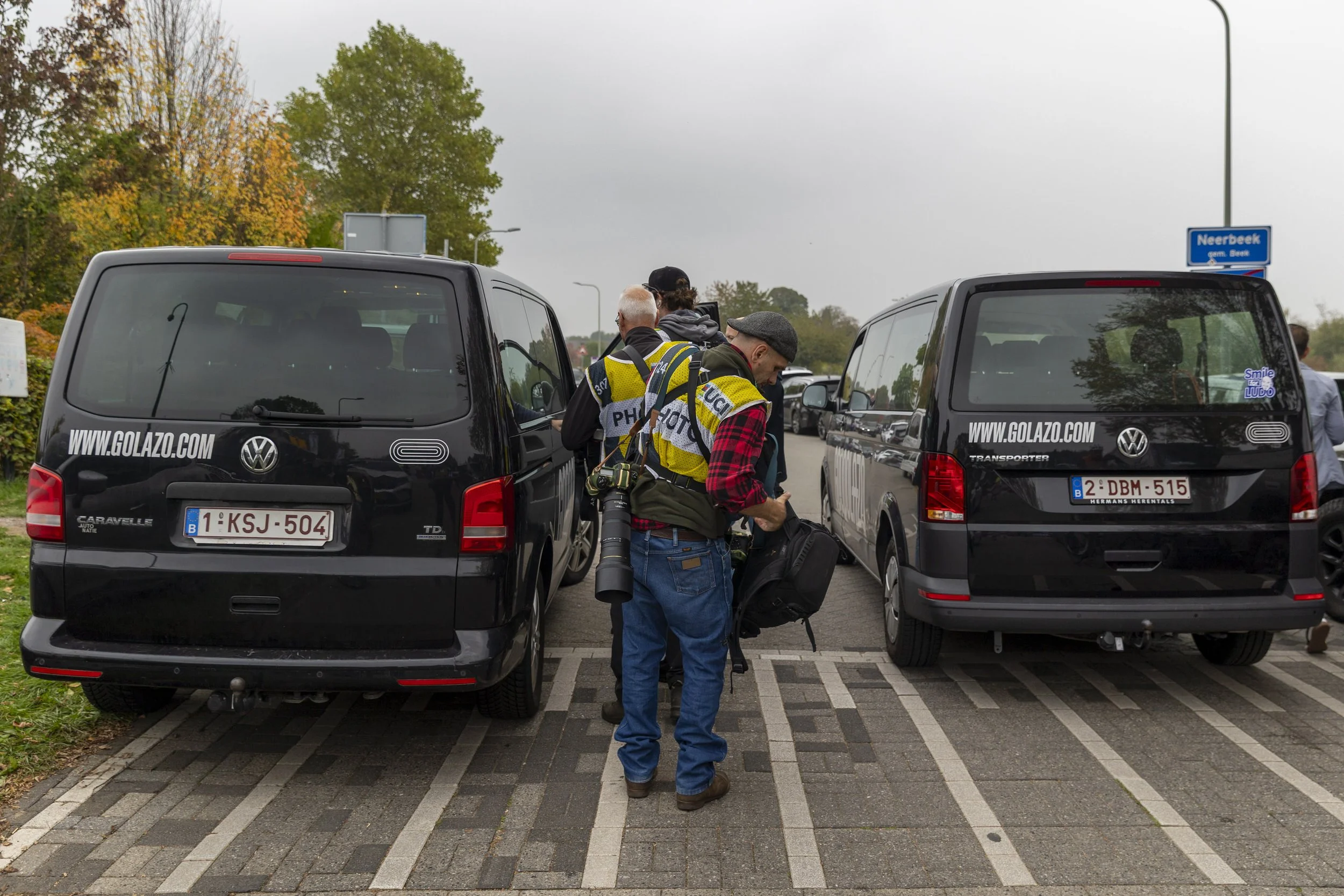 Photographers gathering in a parking lot next to black vans with website URLs and Belgian license plates, with trees in the background and people preparing their photography gear.