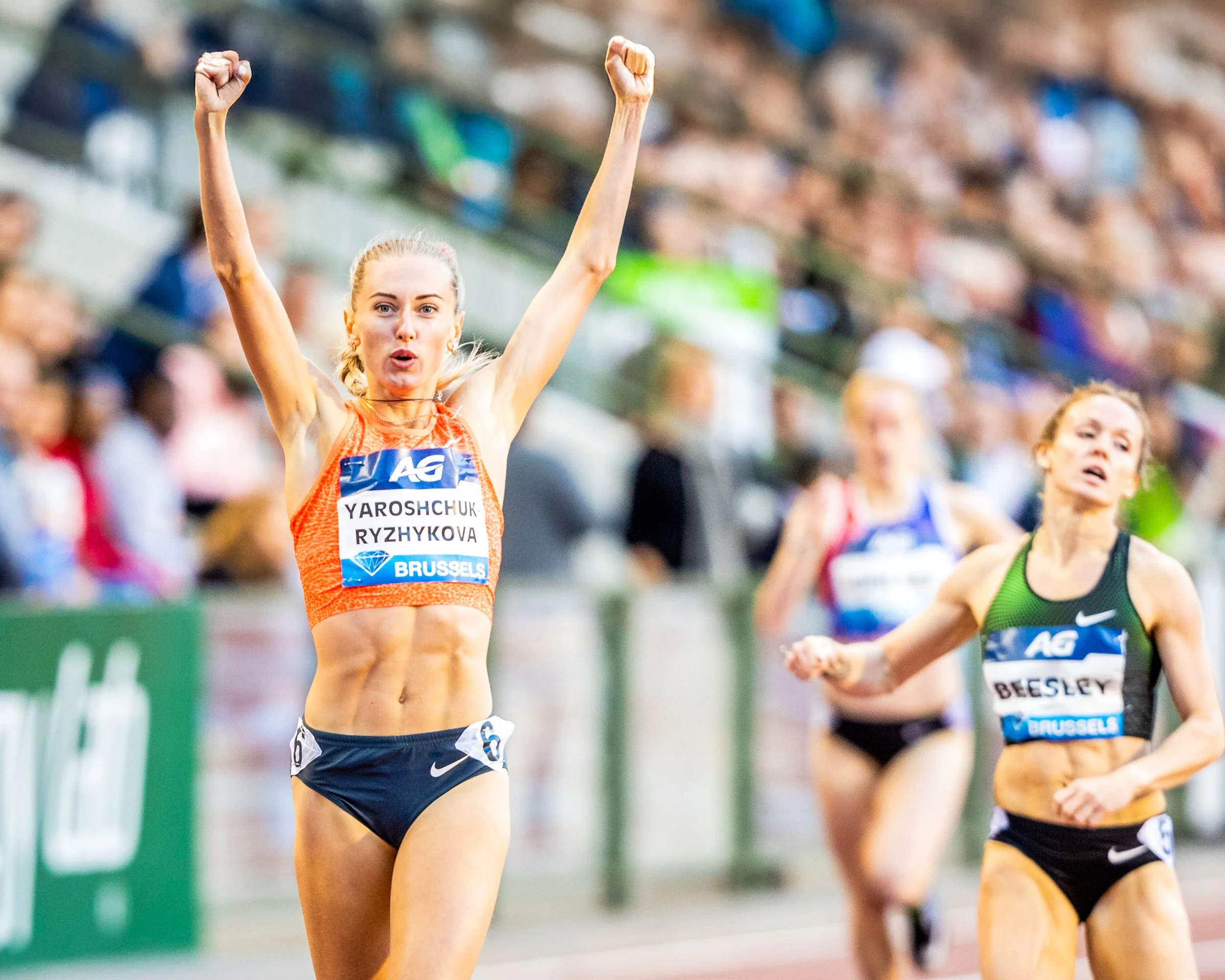 Female athlete celebrating with raised fists during a race, wearing navy shorts, orange top, and a name bib, on an indoor track with other athletes and spectators in the background.