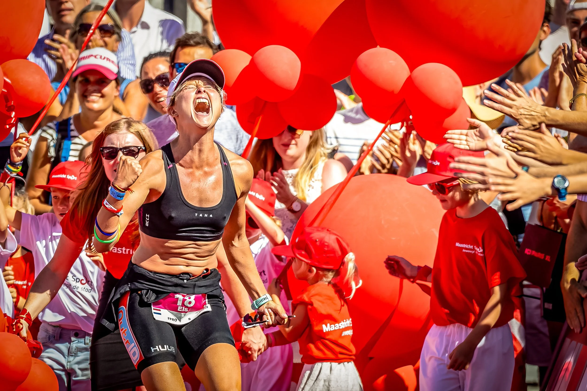 A crowd of people participating in a marathon race, with a woman running in the foreground, wearing a black sports bra and black shorts, surrounded by orange balloons and cheering spectators.