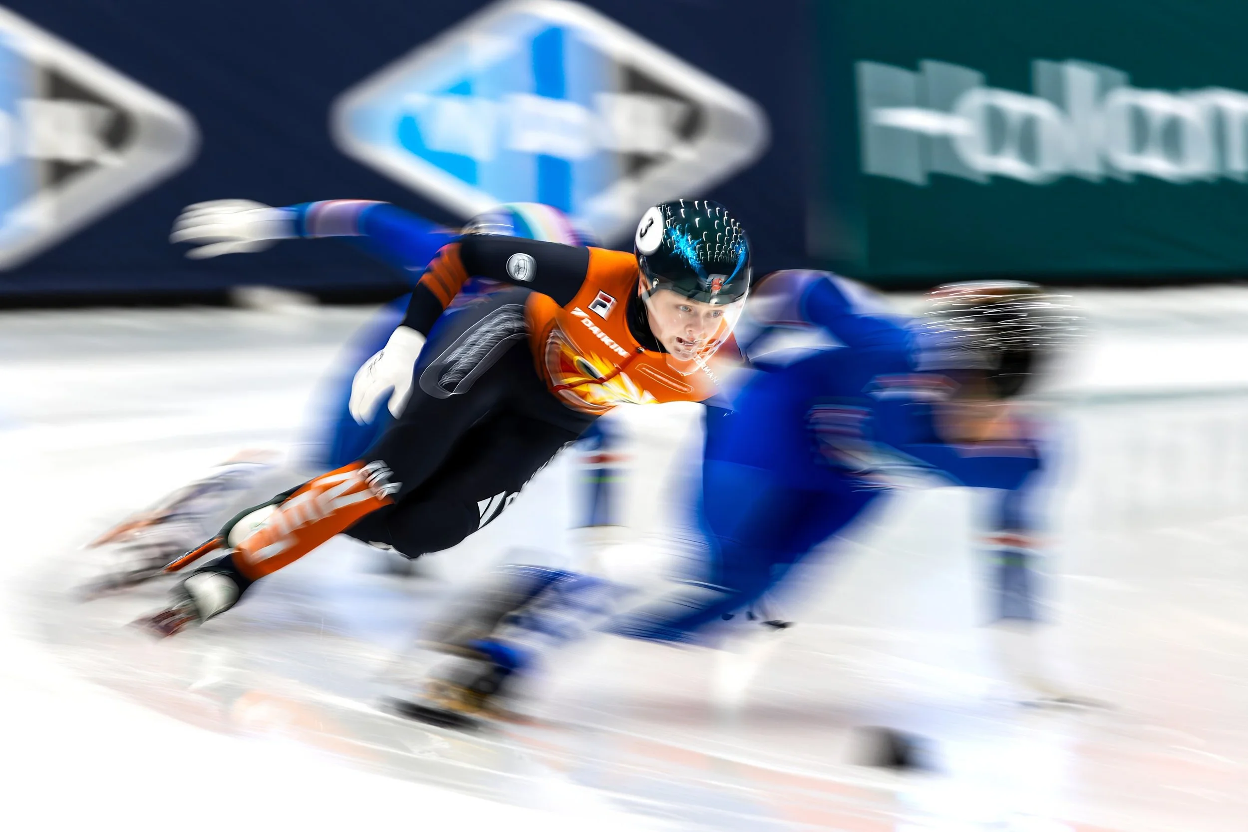 Two speed skaters racing on ice, wearing helmets and colorful uniforms, with motion blur indicating high speed, and a blue and white background.