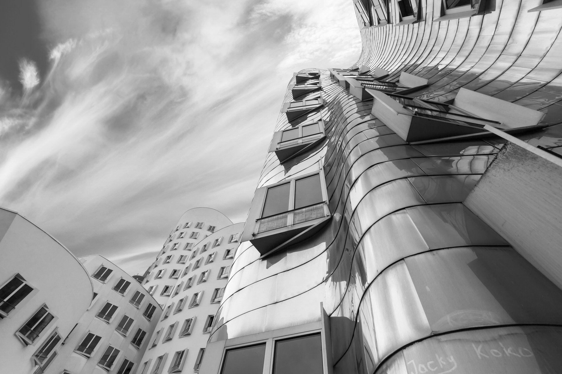 A black and white photo of a modern, curved building with reflective glass windows and a wavy facade, under a cloudy sky.