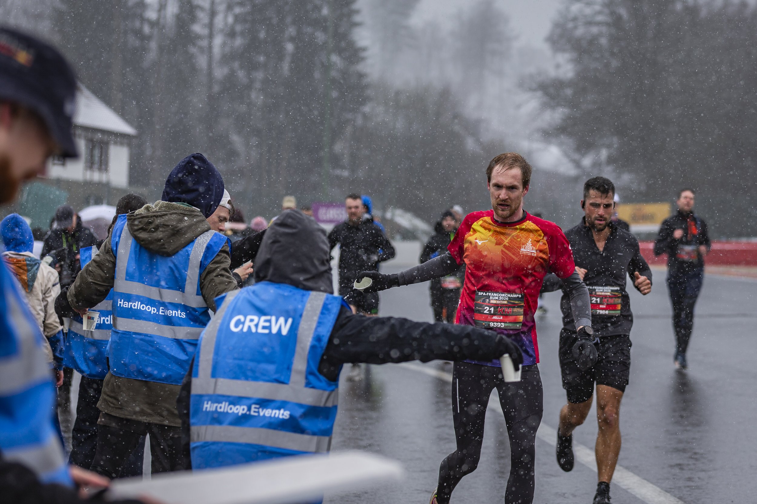 Marathon runners participating in a race on a rainy day, with race bibs, surrounded by event staff and spectators, with trees and buildings in the background.