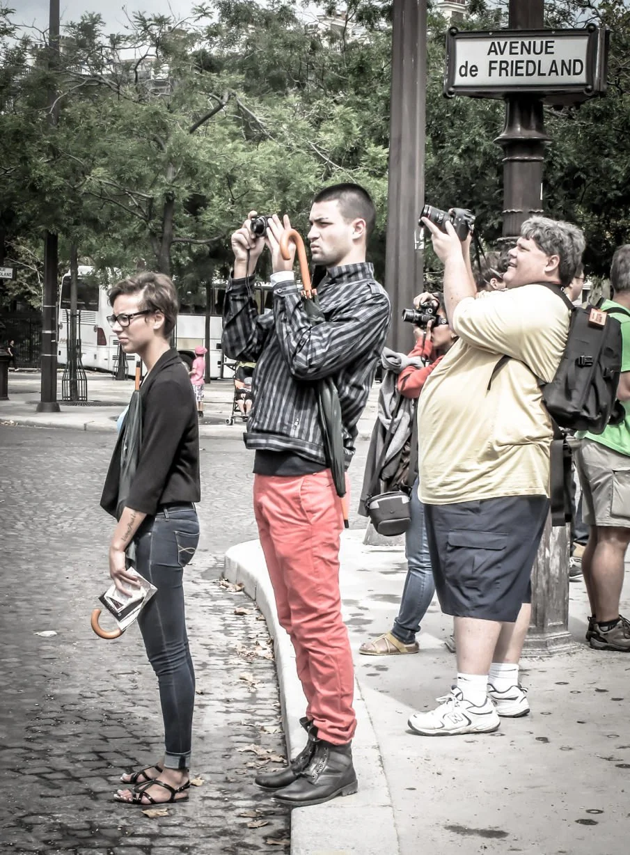 People standing and taking photos on a sidewalk at the intersection of Avenue de Friedland with trees and a bus in the background.